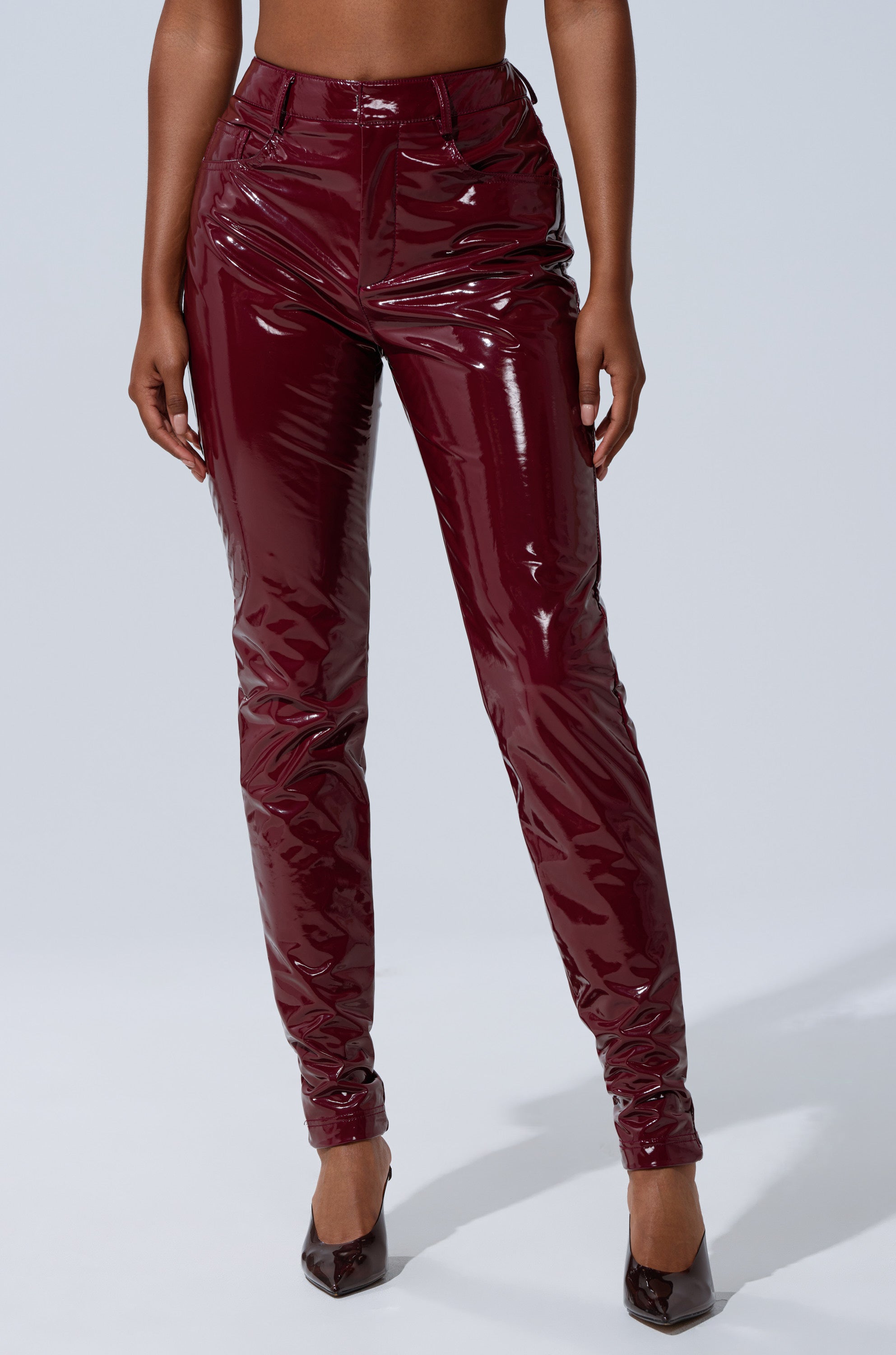 A person models the ON FIRE FAUX LEATHER LEGGING—high-rise, skinny, burgundy faux leather pants—paired with pointed high heels against a light gray background, showing only the lower torso and legs.