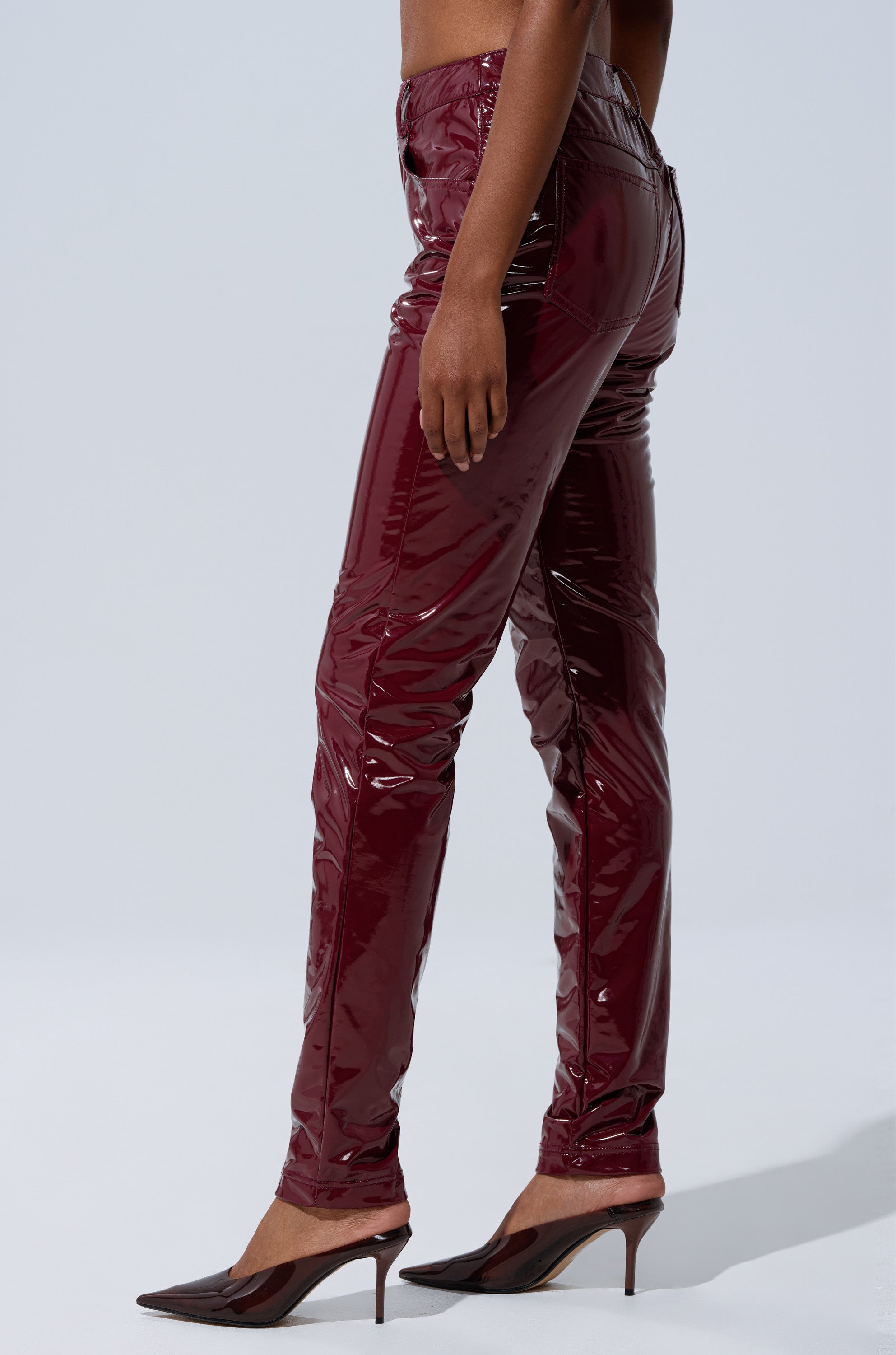 Wearing the ON FIRE FAUX LEATHER LEGGING in shiny, high-rise burgundy, paired with pointed-toe burgundy heels, a person stands against a plain light background, shown from the waist down in a side view.