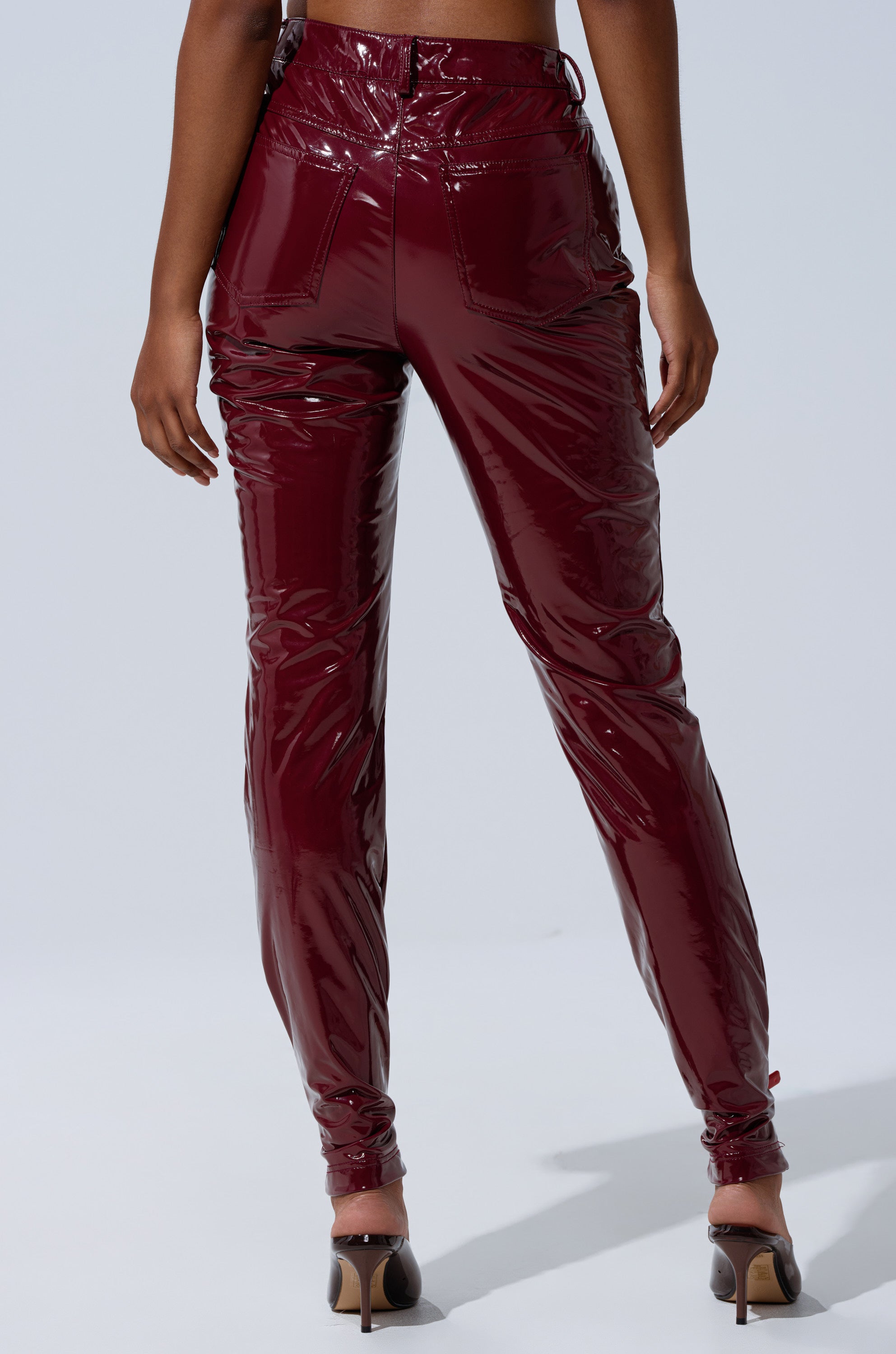 A person stands facing away, wearing ON FIRE FAUX LEATHER LEGGING in burgundy with a high-rise skinny fit and shiny vinyl finish, styled with open-toe heels against a plain light background.