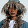 Dressed in a SADE FUR TRIM DENIM BUCKET HAT and a jacket with fluffy, textured material, an individual exudes style. The hat is distinguished by its wide faux fur brim, while the jacket showcases faux fur accents on the shoulders, enhancing their accessory game. They gaze directly at the camera against a plain background.