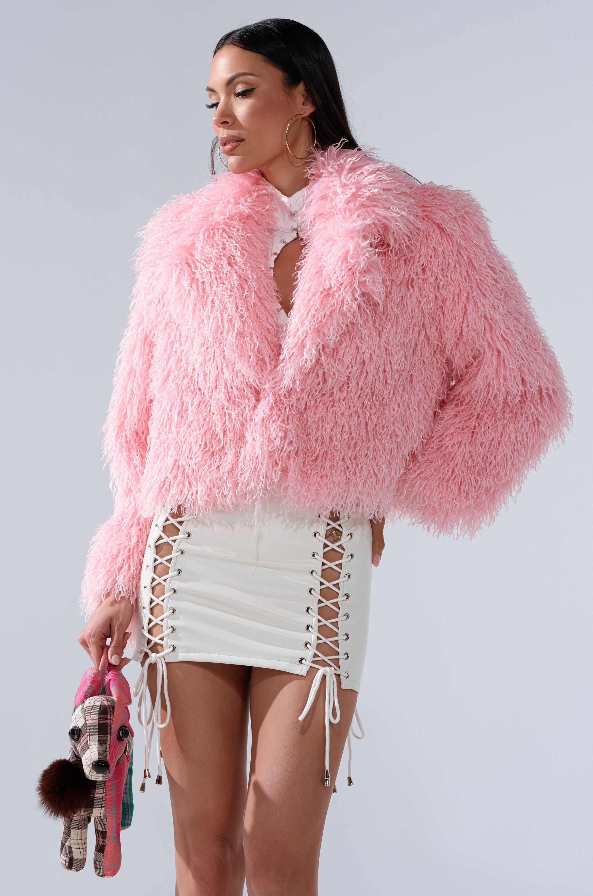 A woman with long dark hair and large hoop earrings wears the DIVINE FEMININE FAUX FUR COAT in plush pink and a short white skirt with lace-up sides, holding a plaid stuffed dog toy decorated with a pom-pom keychain.