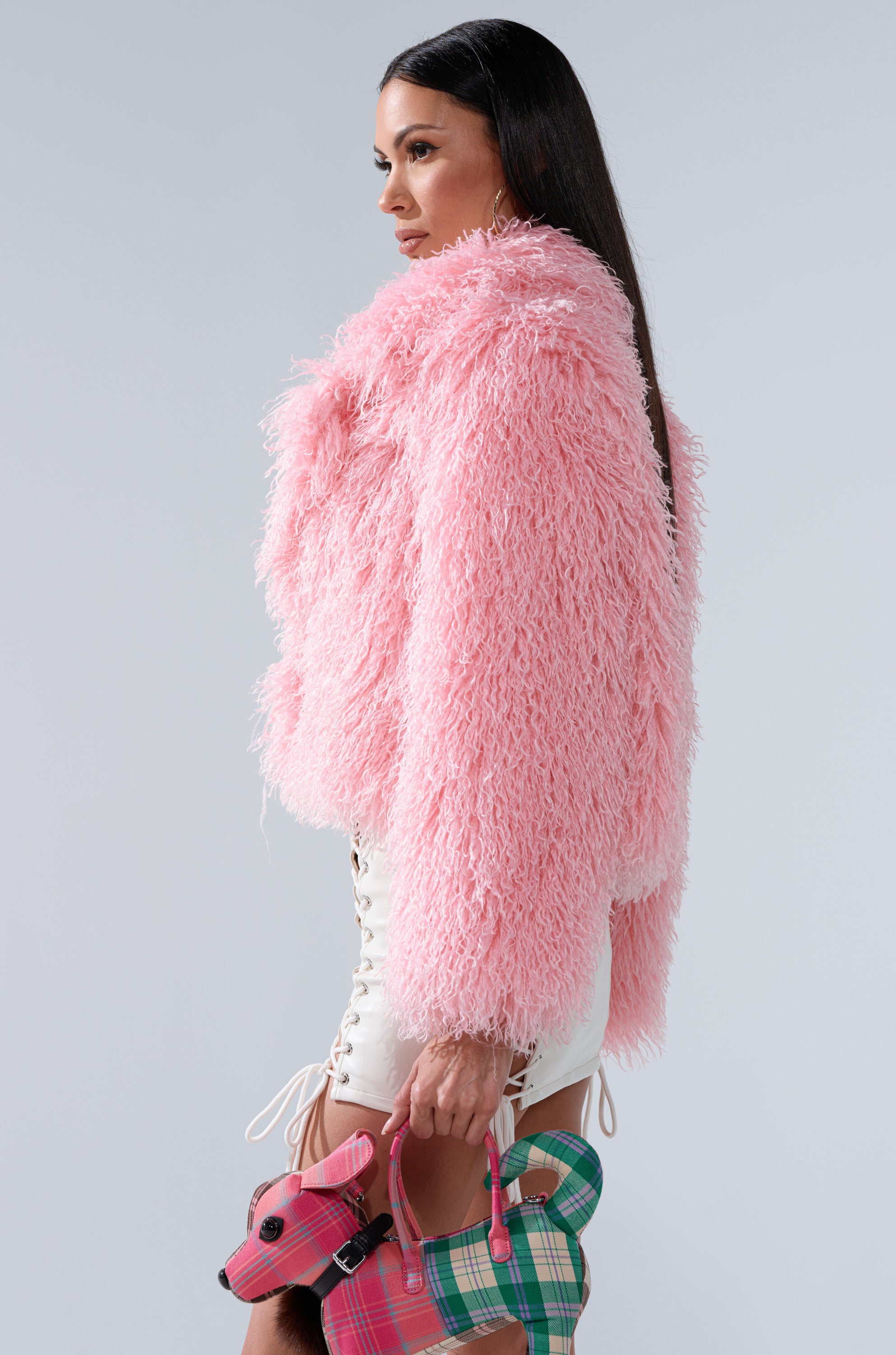 Wearing the DIVINE FEMININE FAUX FUR COAT in plush pink, a woman pairs it with white lace-up shorts and holds a small plaid dog-shaped handbag against a plain light gray background.