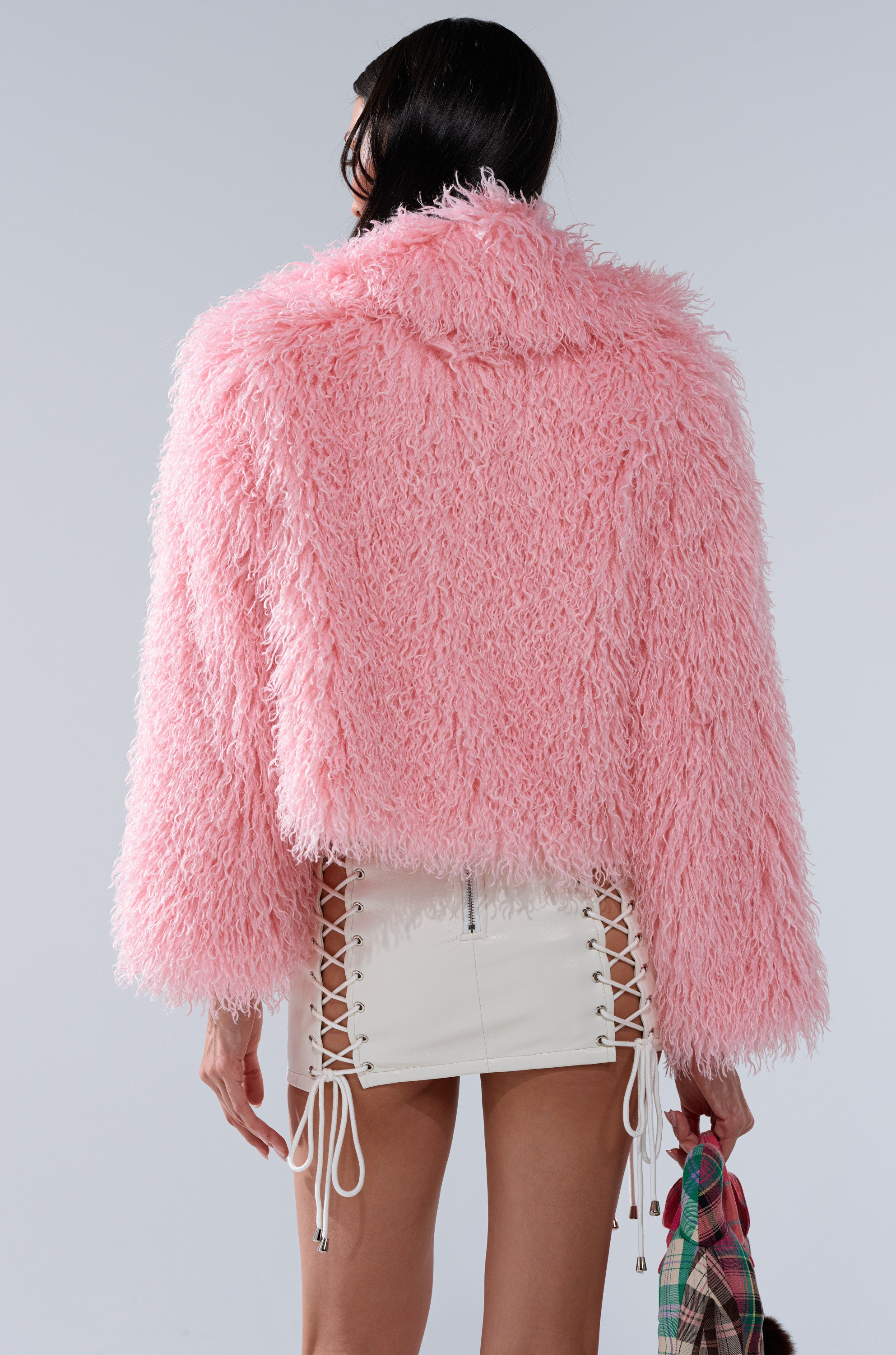 A person with long dark hair is shown from the back wearing the DIVINE FEMININE FAUX FUR COAT in plush pink and a white mini skirt with lace-up sides, holding a plaid item.