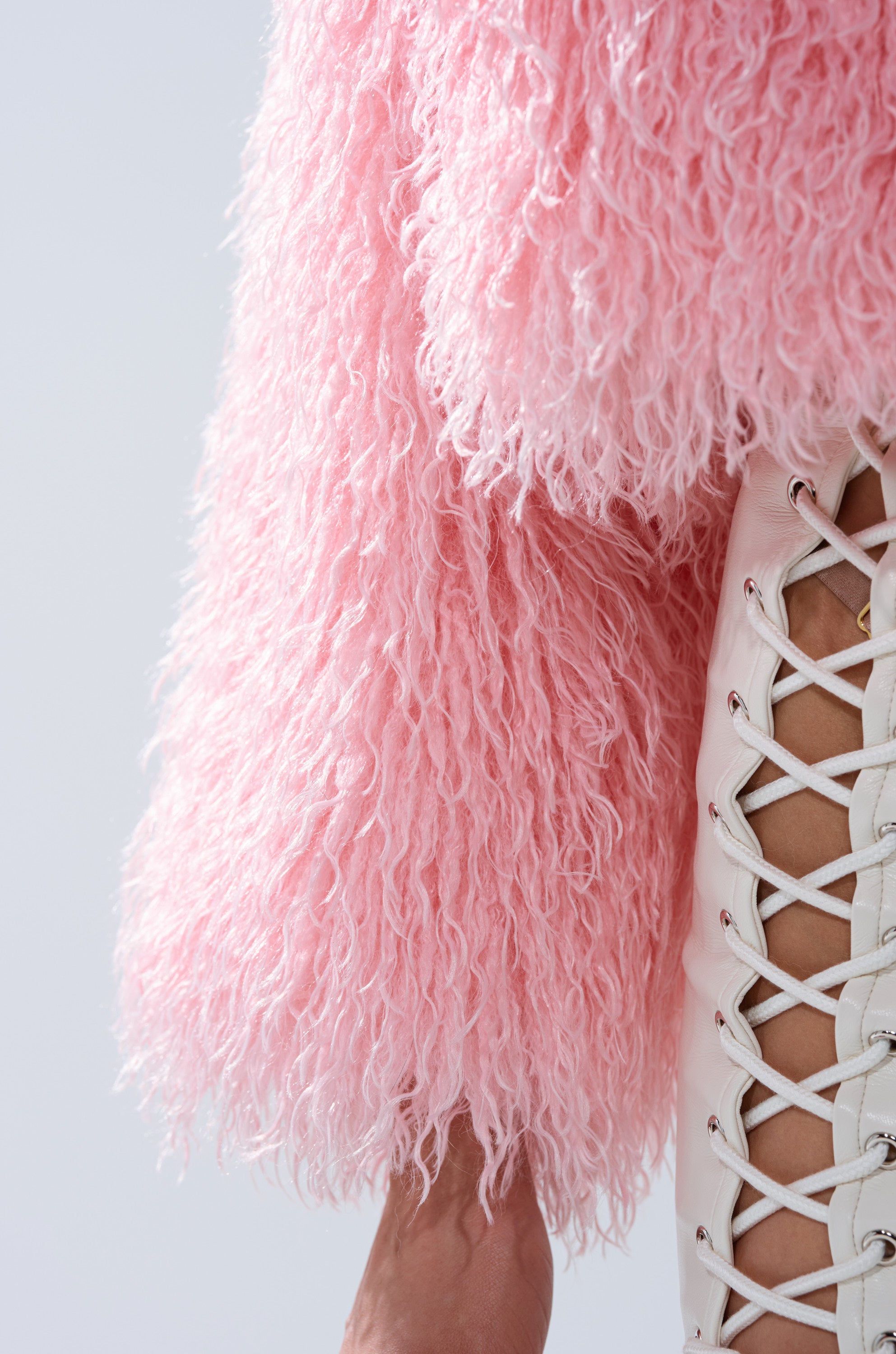 A person is seen in a close-up shot wearing the DIVINE FEMININE FAUX FUR COAT layered over a textured pink sweater and a white lace-up top, with exposed skin, against a light background for added style.