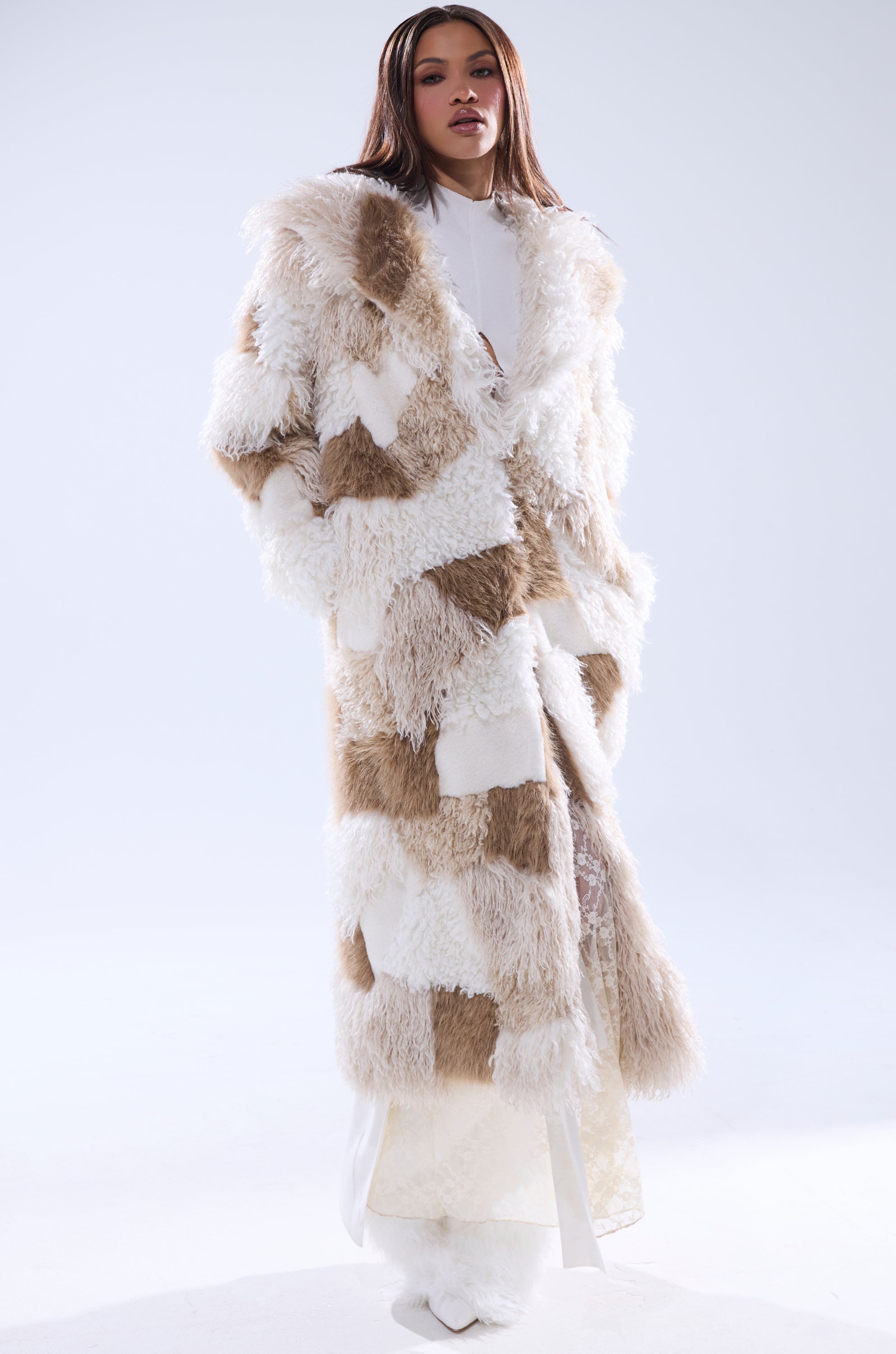 A woman with straight brown hair and a confident look wears the IN MY FEELS PATCHWORK FAUX FUR COAT in white and beige over a white outfit, standing against a plain light background.