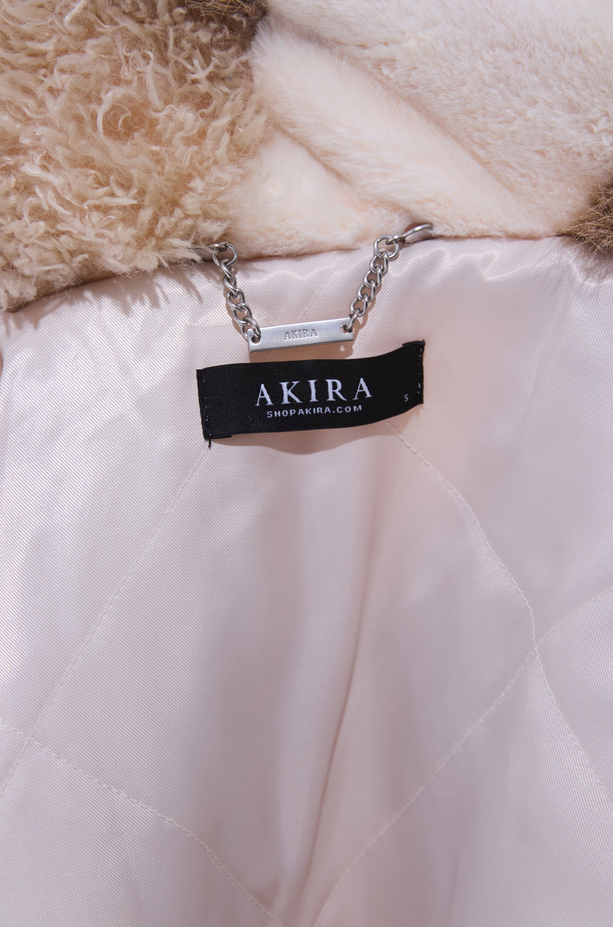 Close-up of the inside of the IN MY FEELS PATCHWORK FAUX FUR COAT, featuring a visible "AKIRA" brand label, a metal chain hanging loop, and a smooth, light-colored inner lining.