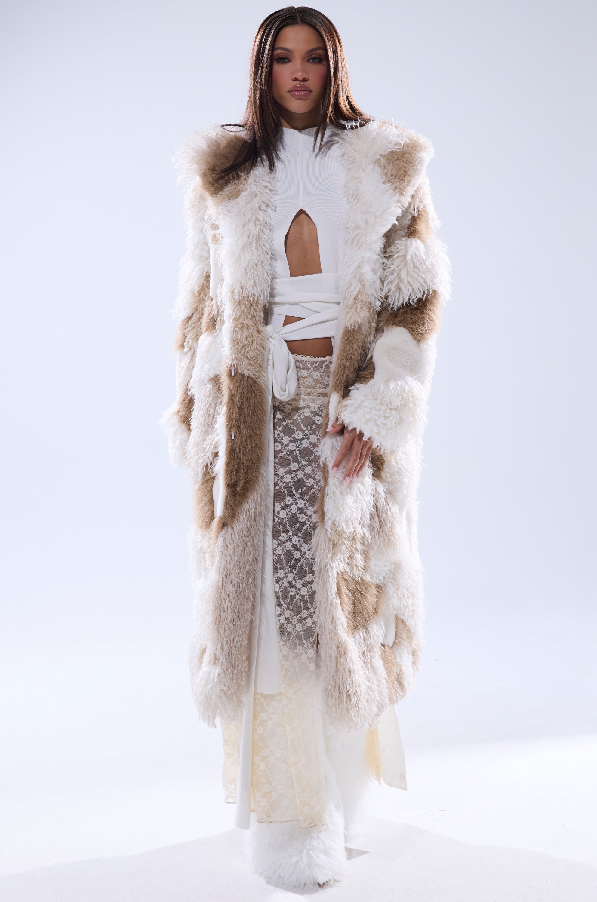 A woman stands confidently in the IN MY FEELS PATCHWORK FAUX FUR COAT, featuring tan and white faux fur, over a white cutout top, sheer lace skirt, and white heels against a plain white background.