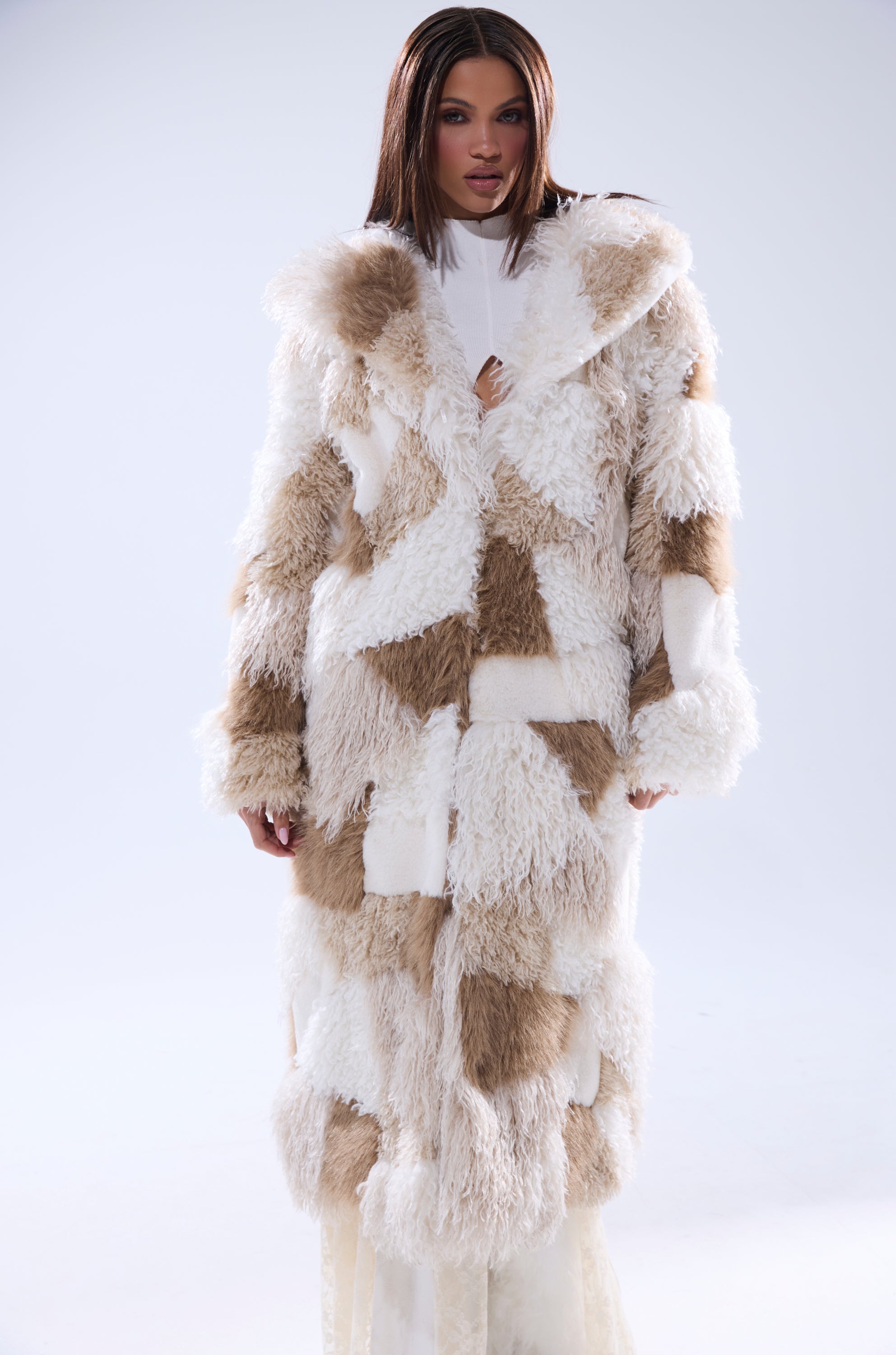 A woman faces forward in the IN MY FEELS PATCHWORK FAUX FUR COAT, featuring long, fluffy white and beige patchwork over a white outfit, set against a plain light background.