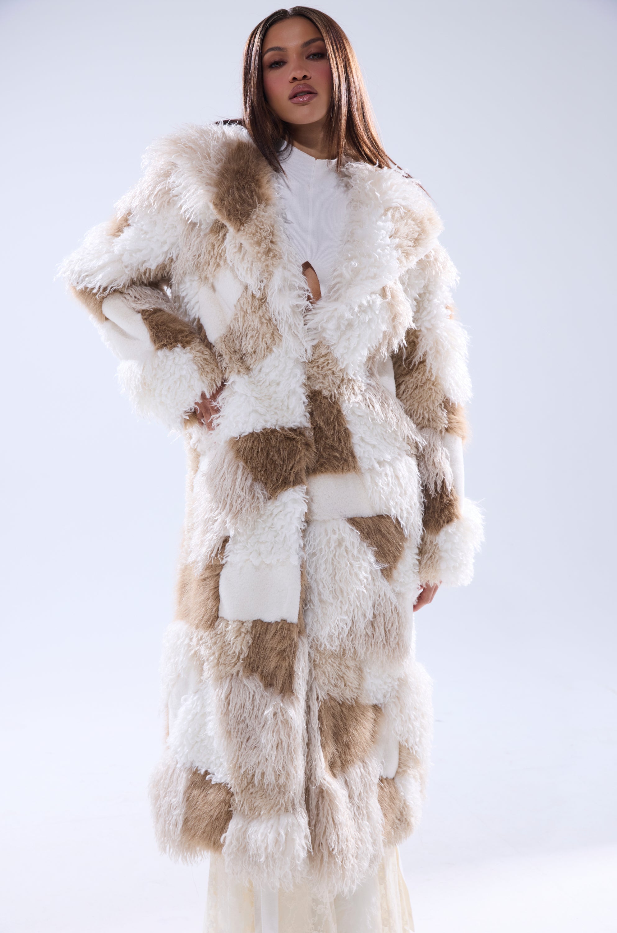 A woman stands confidently in the IN MY FEELS PATCHWORK FAUX FUR COAT, featuring beige and white tones over a white outfit, set against a plain light background.