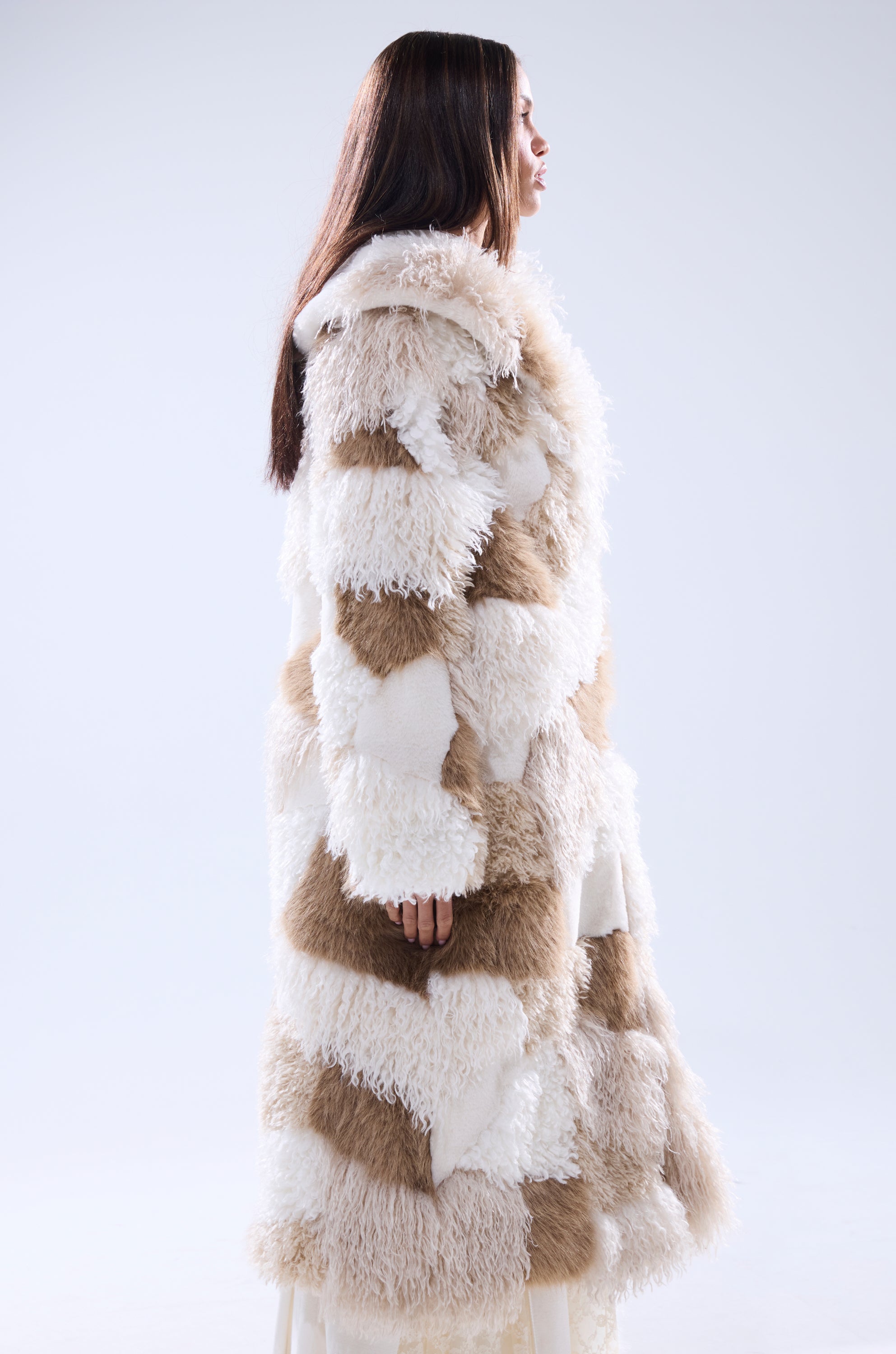A woman with long brown hair stands in profile, wearing the IN MY FEELS PATCHWORK FAUX FUR COAT in cream and tan against a plain light background.