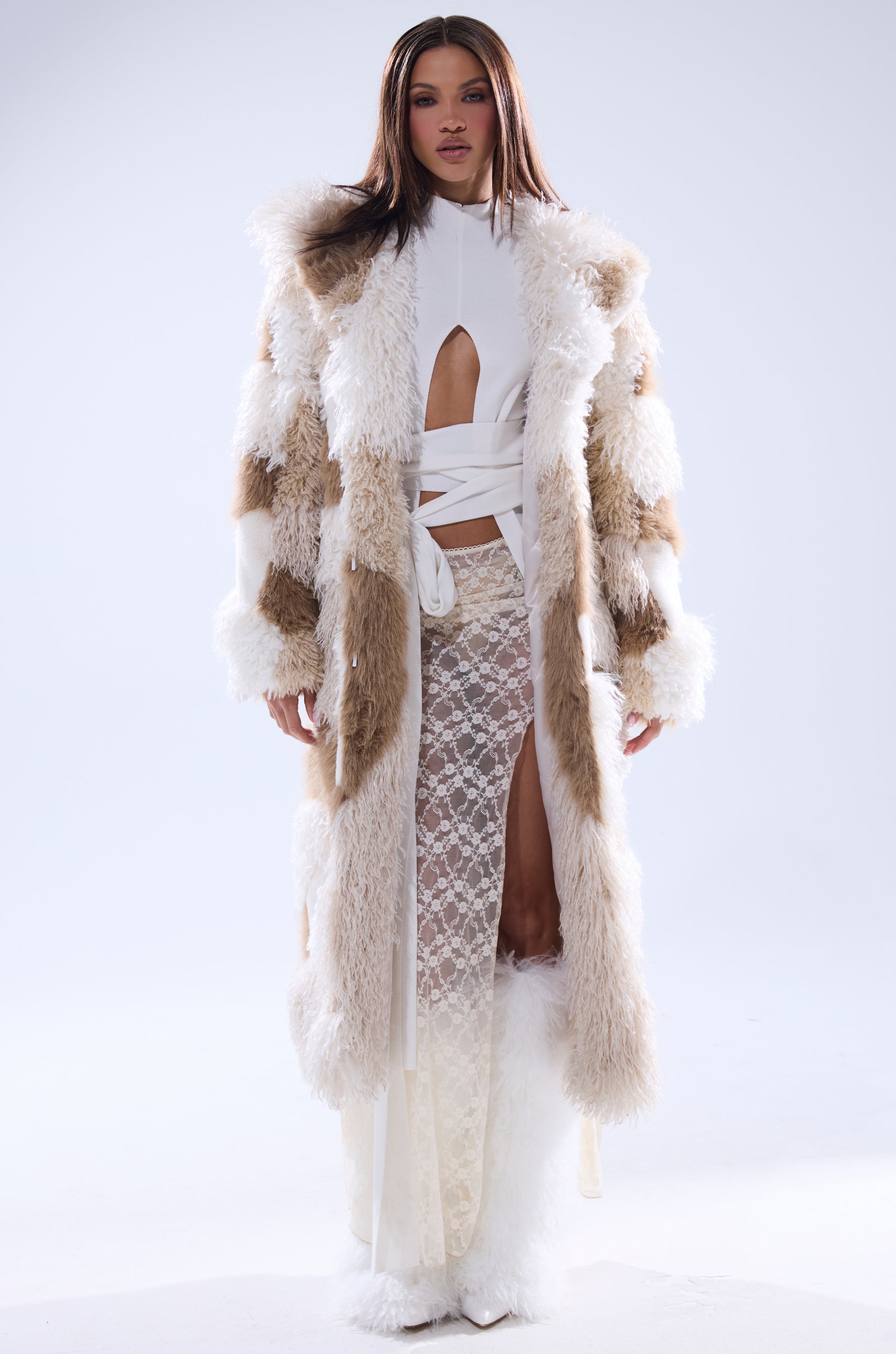A woman stands confidently in the IN MY FEELS PATCHWORK FAUX FUR COAT, featuring beige and white faux fur, styled over a white cut-out top and sheer lace high-slit skirt against a plain, light background.