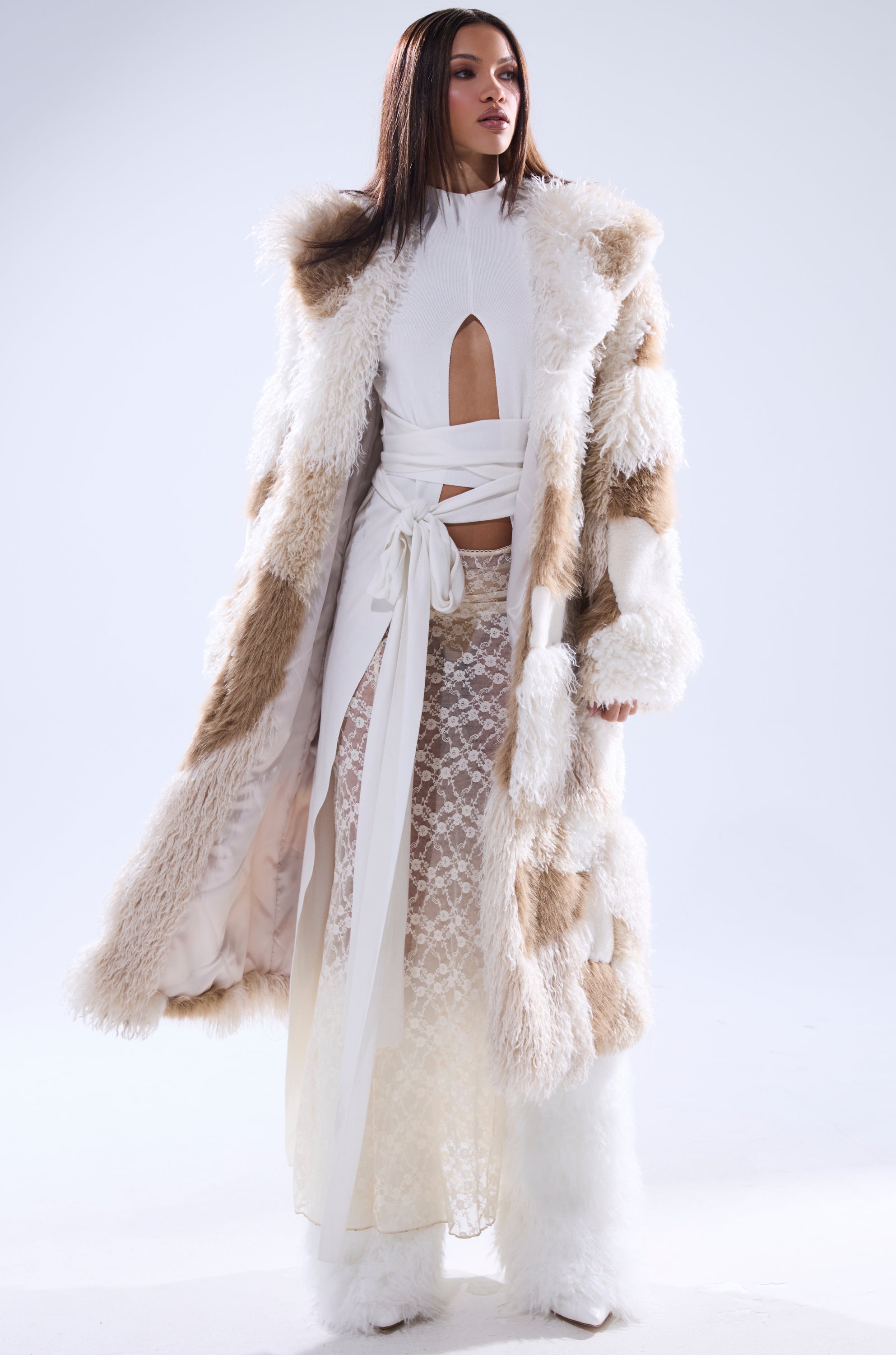 A woman stands confidently in a studio with soft lighting, wearing the IN MY FEELS PATCHWORK FAUX FUR COAT—long and luxurious with beige and white patches—over a white cutout dress featuring a waist tie and sheer lace skirt.