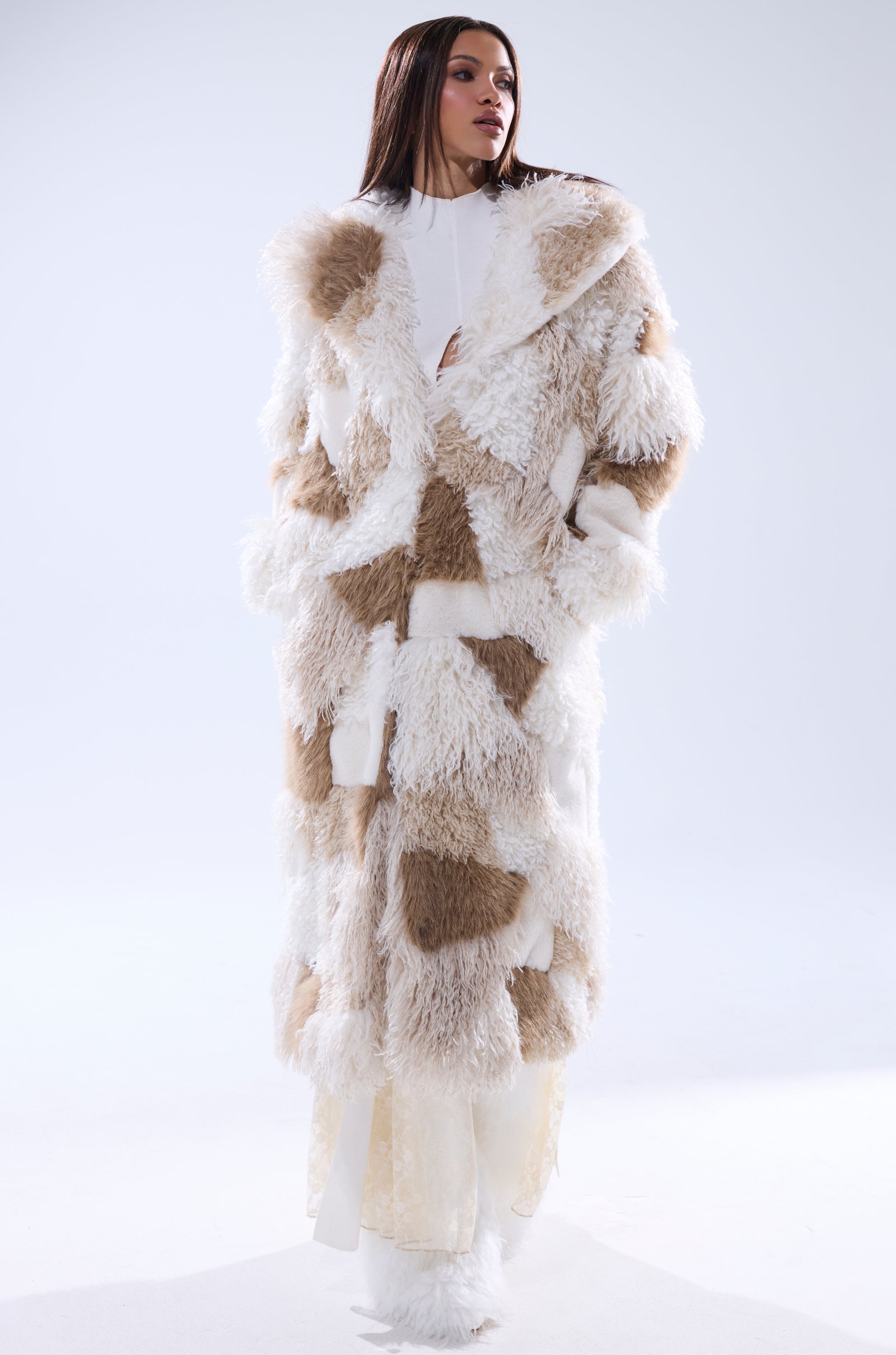 A person wears the IN MY FEELS PATCHWORK FAUX FUR COAT, featuring a long, fluffy texture with white and light brown patches, standing against a plain light background and looking slightly to the side with hands in pockets.
