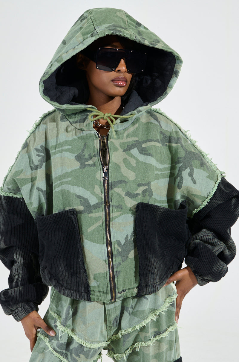 A person is styled in a green, classic camo print oversized hoodie and pants featuring black accents. The hoodie includes a large hood and frayed edges, paired perfectly with the padded quilted inner lining of the AKIRA Label CADET MIXED MEDIA BOMBER. They complete their unique, casual look with large, dark sunglasses.