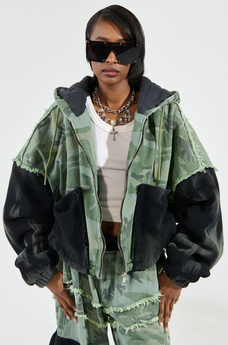 A person is standing against a plain white background, wearing oversized black sunglasses and the green CADET MIXED MEDIA BOMBER featuring a classic camo print with black sleeves, paired with matching camouflage pants. The individual has short hair.