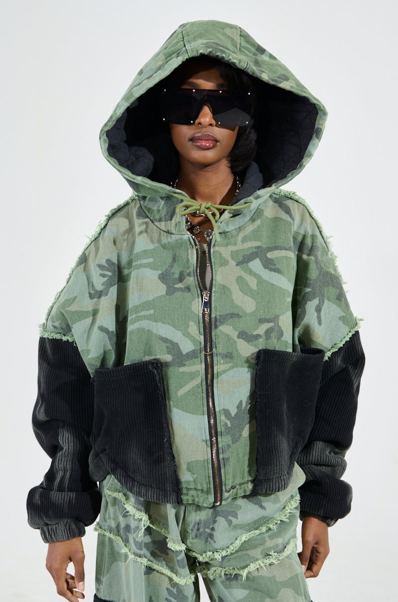 A person stands against a plain background, wearing matching pants and sporting the CADET MIXED MEDIA BOMBER with its classic camo print jacket and hood up. The look is completed with large, dark sunglasses that pair perfectly while concealing a padded quilted inner lining within this stylish attire.