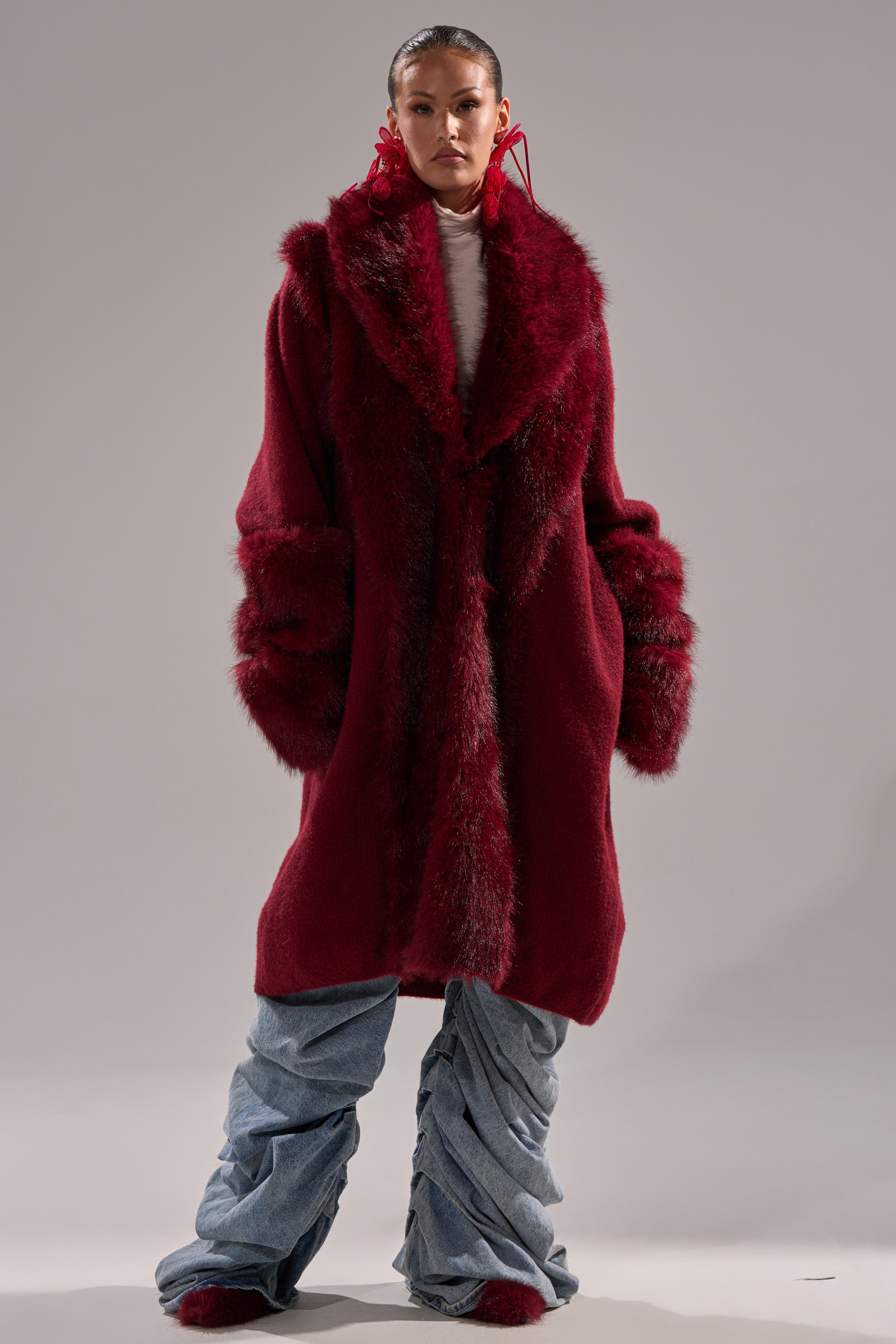 A person wears the LUXE LIFE FUR CARDIGAN IN BURGUNDY, styled over light blue ruched jeans and red shoes with bold red earrings, standing against a plain gray background.