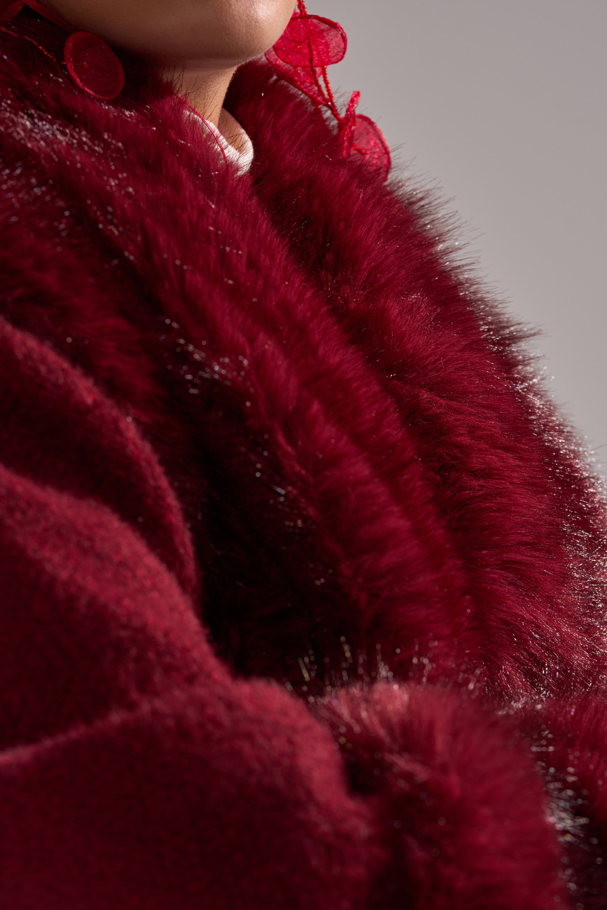 A person models the LUXE LIFE FUR CARDIGAN IN BURGUNDY—a deep red faux fur coat—paired with red earrings, showing only the lower face and neck against a neutral background.
