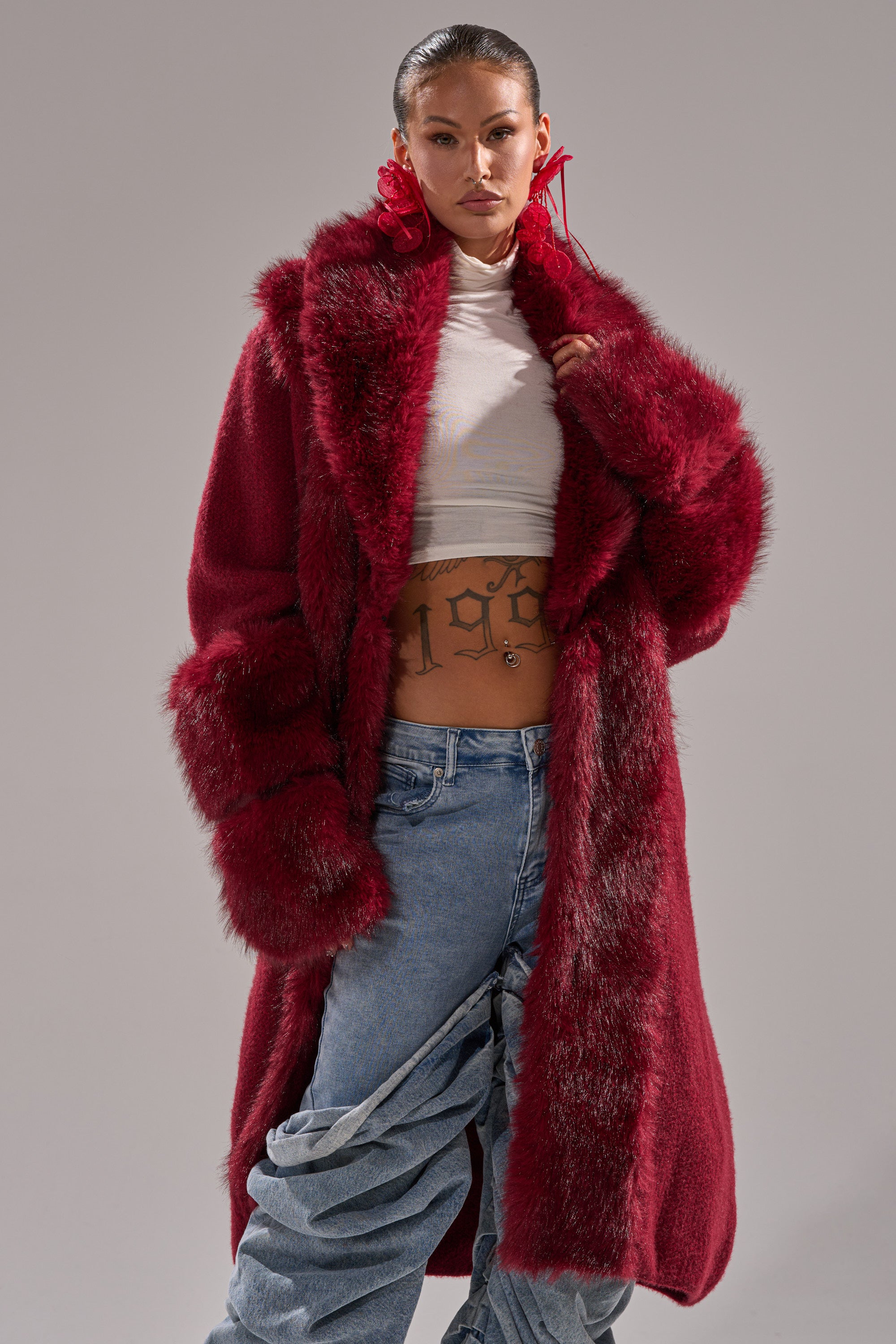 A woman models the Luxe Life Fur Cardigan in Burgundy, styled over a white cropped turtleneck that shows her "1991" stomach tattoo. She completes the look with ruched blue jeans and oversized red earrings for standout style.
