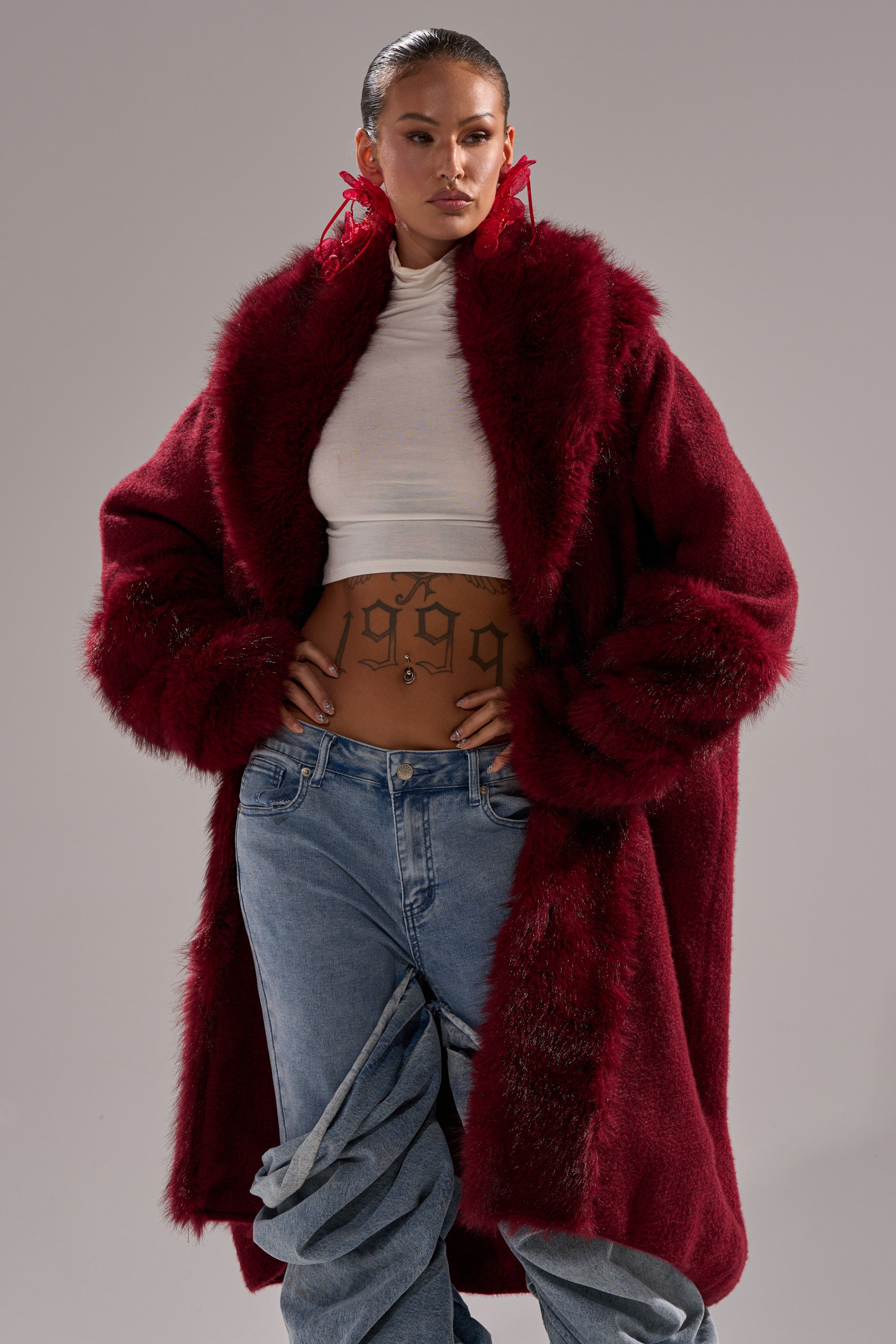 A woman wears a LUXE LIFE FUR CARDIGAN IN BURGUNDY over a white turtleneck crop top and loose blue jeans, accessorized with red earrings, a belly button piercing, and a "1999" stomach tattoo.