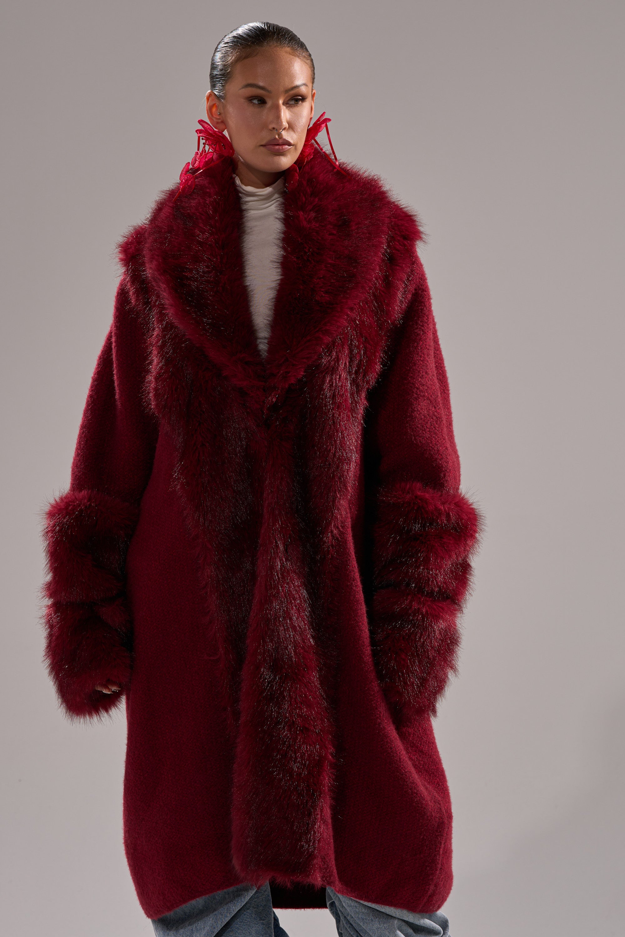 Someone poses against a plain gray background wearing the LUXE LIFE FUR CARDIGAN IN BURGUNDY, featuring a large collar and cuffs, layered over a white turtleneck.