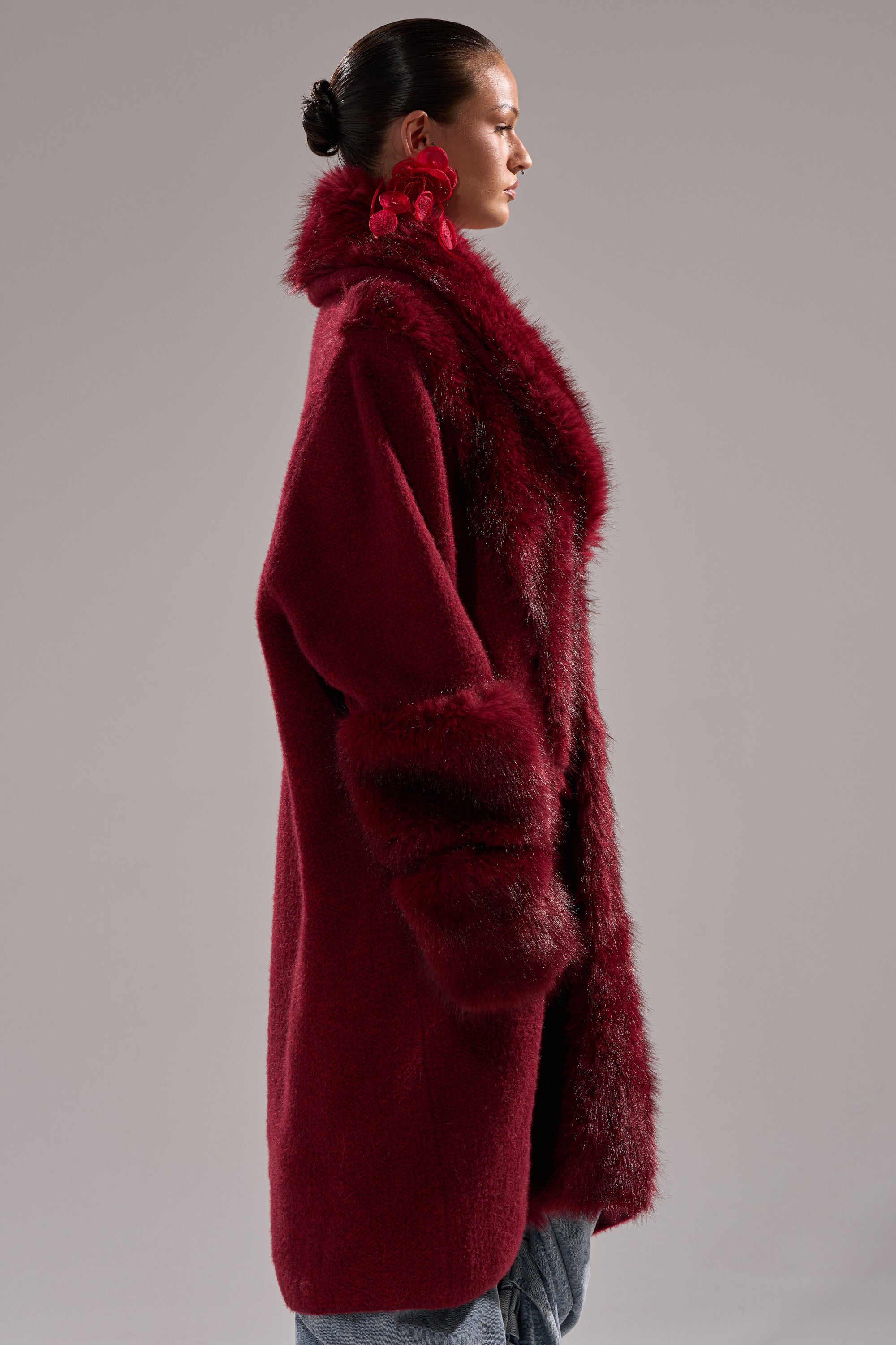 A woman with her hair in a sleek bun wears the LUXE LIFE FUR CARDIGAN IN BURGUNDY—long and plush with fur trim—and large red earrings, standing against a plain light background.