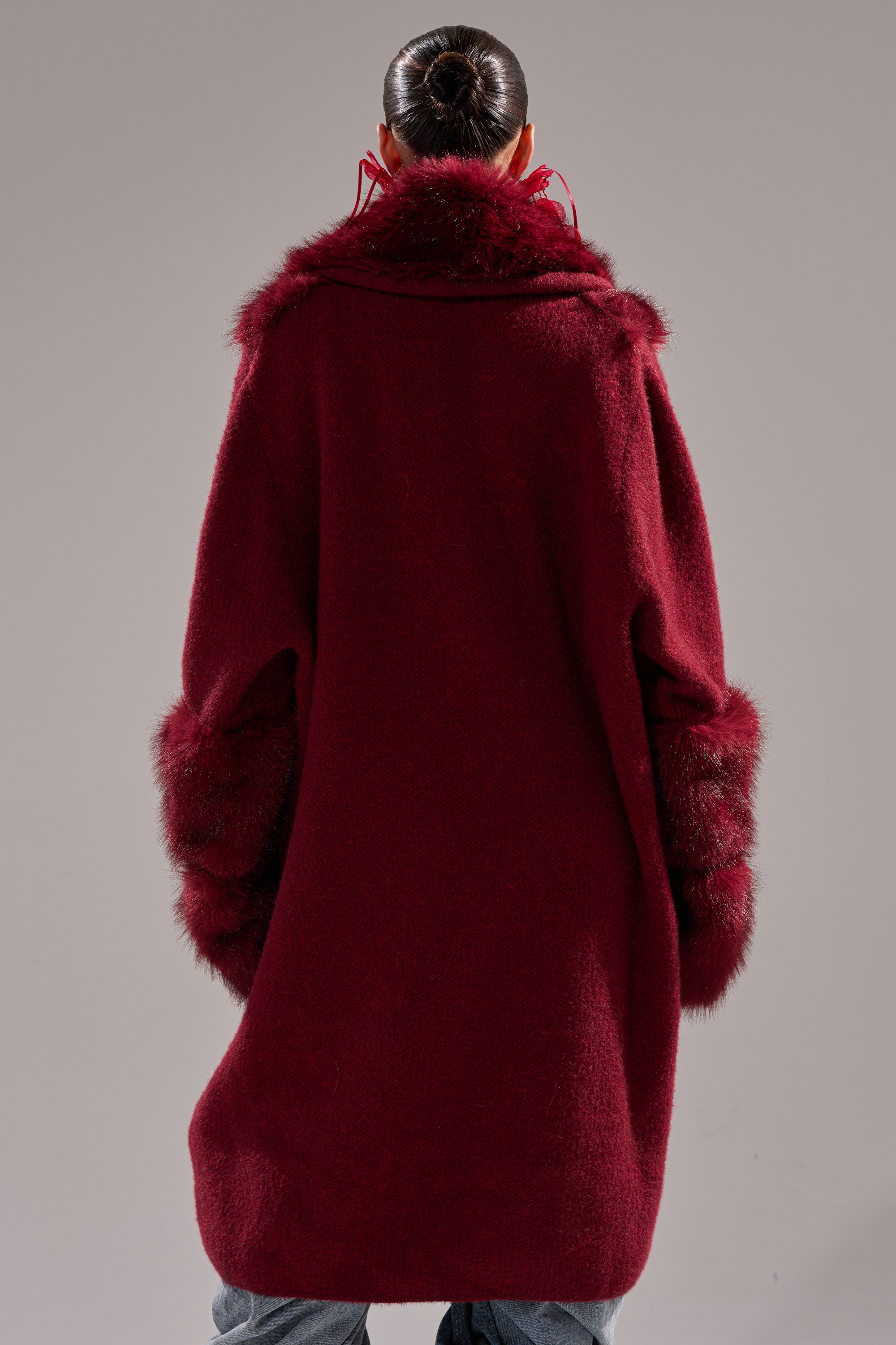 A person with a sleek bun faces away from the camera, wearing the LUXE LIFE FUR CARDIGAN IN BURGUNDY—a long, deep red faux fur cardigan with fur trim on the collar and cuffs—against a plain background.