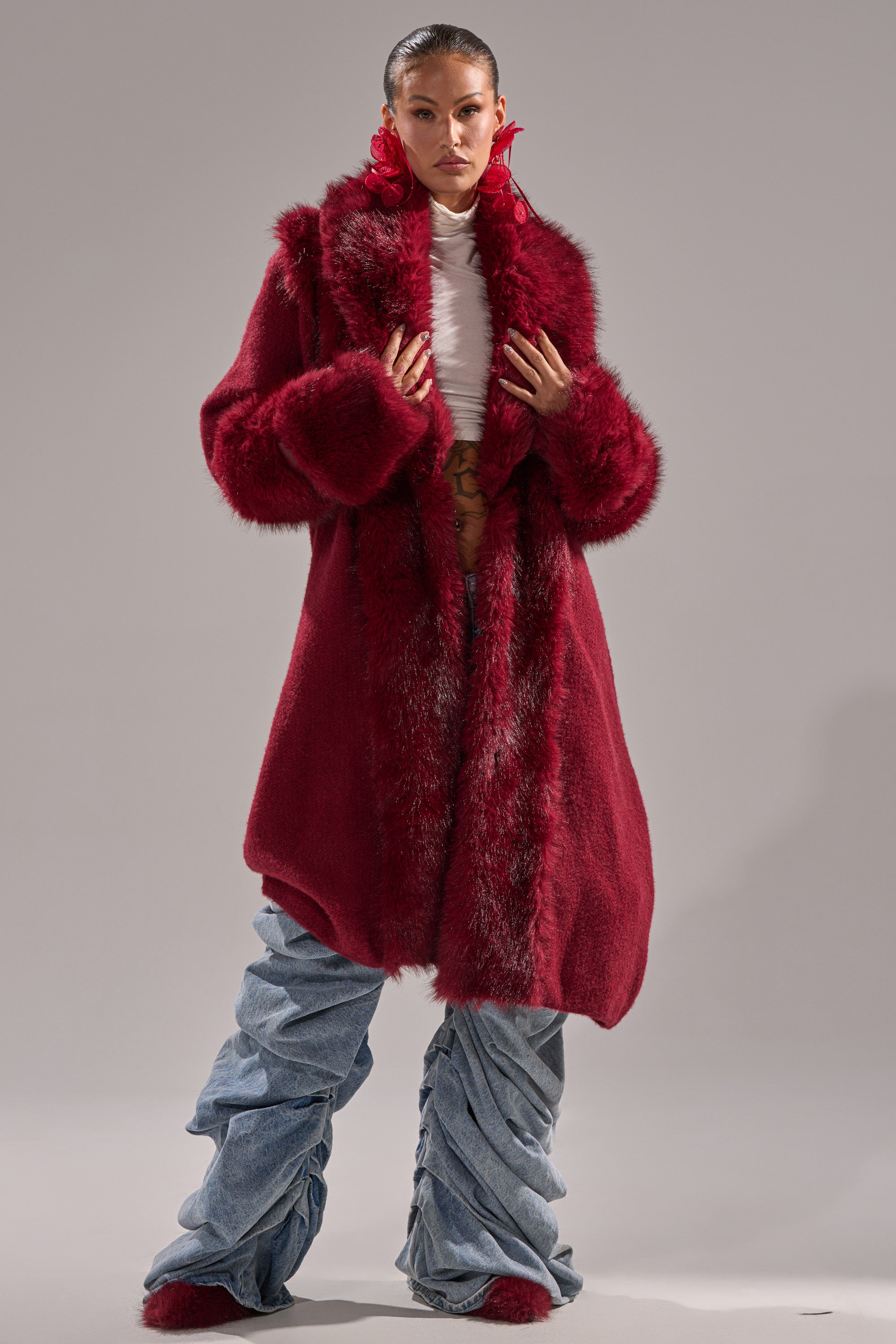 A person stands confidently in the LUXE LIFE FUR CARDIGAN IN BURGUNDY over a white top and ruched light blue jeans, accessorized with bold red earrings against a plain gray background.