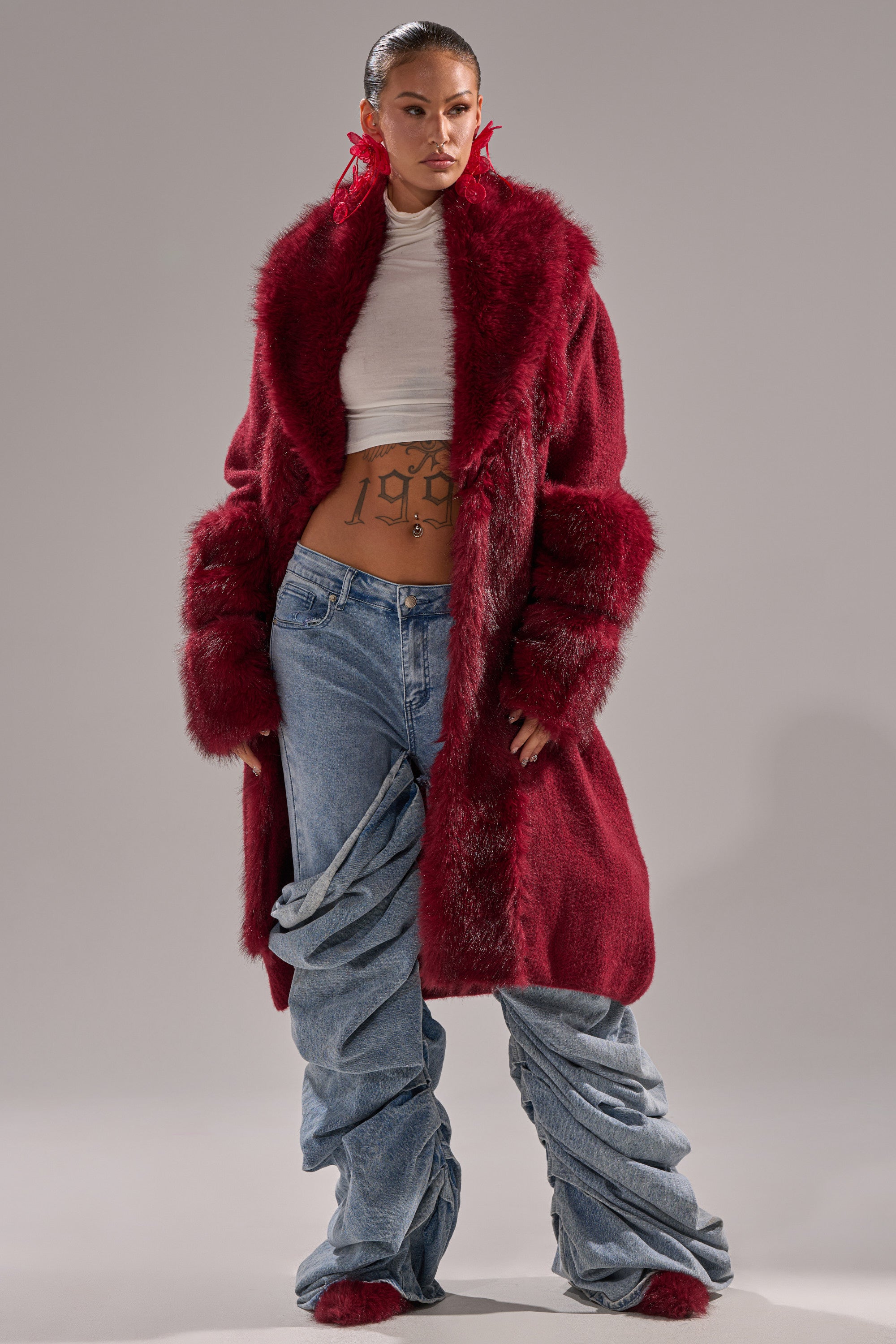 A woman poses in the LUXE LIFE FUR CARDIGAN IN BURGUNDY, a white turtleneck crop top, and loose-fitting ruched blue jeans. Her hair is slicked back, she wears bold red earrings, and a "1998" tattoo is visible on her midriff.