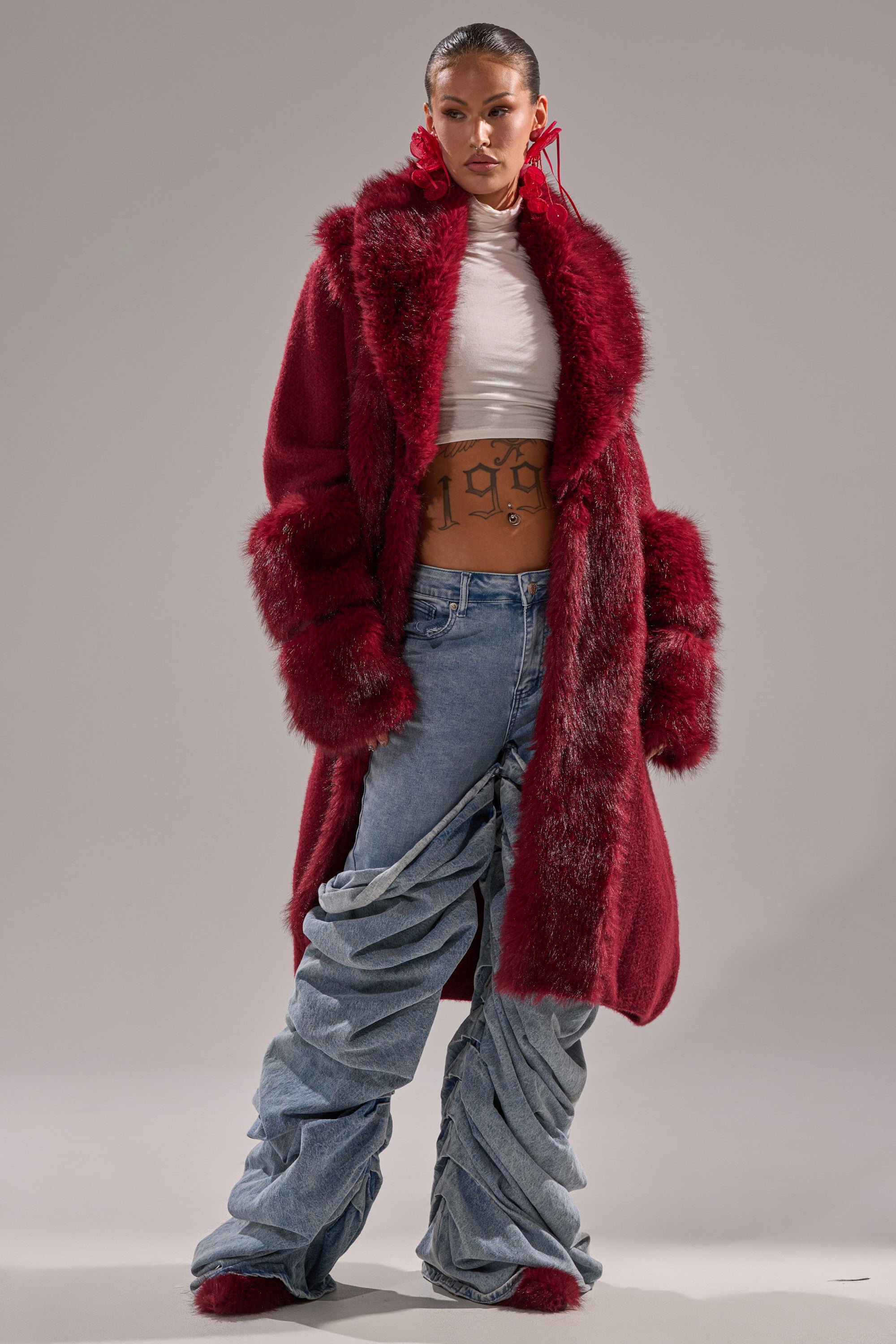 A person stands confidently in the LUXE LIFE FUR CARDIGAN IN BURGUNDY, paired with a white crop top, ruched blue jeans, and bold red earrings, highlighting a striking and modern fashion look against a plain background.