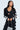 A woman confidently poses in a black FEELING CUTE BOW DETAIL BLAZER from AKIRA Label, featuring white sleeve and pant bows. She pairs it with a black bralette, silver handbag, loose long hair, and hoop earrings.