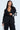 A fashionable woman poses in the AKIRA Label FEELING CUTE BOW DETAIL BLAZER, featuring white bows on a black top. She holds a silver handbag, with long dark hair cascading over her shoulders, creating a confident and stylish look.