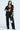 A woman poses in a sleek black FEELING CUTE BOW DETAIL BLAZER, featuring white bows on the sleeves and pants. She pairs it with a black crop top and sparkles with a silver handbag, her long hair down against a light backdrop, exuding elegance and confidence.