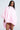 A woman with long straight hair, glasses, and a neutral expression confidently models the FLOATING NYLON BUBBLE BOMBER IN LIGHT PINK—a voluminous, bomber-inspired cape dress with puffed sleeves and a high collar—against a plain background.