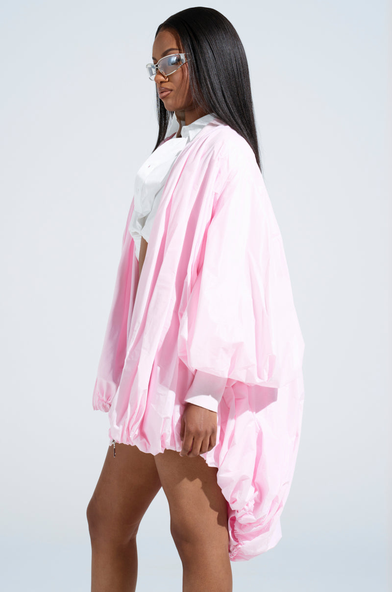 A woman with long straight hair wears the FLOATING NYLON BUBBLE BOMBER IN LIGHT PINK, a white shirt, and clear visor sunglasses. She stands sideways against a plain background, embodying effortless streetwear with a neutral expression.