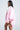 A woman with long straight hair wears the FLOATING NYLON BUBBLE BOMBER IN LIGHT PINK, a white shirt, and clear visor sunglasses. She stands sideways against a plain background, embodying effortless streetwear with a neutral expression.