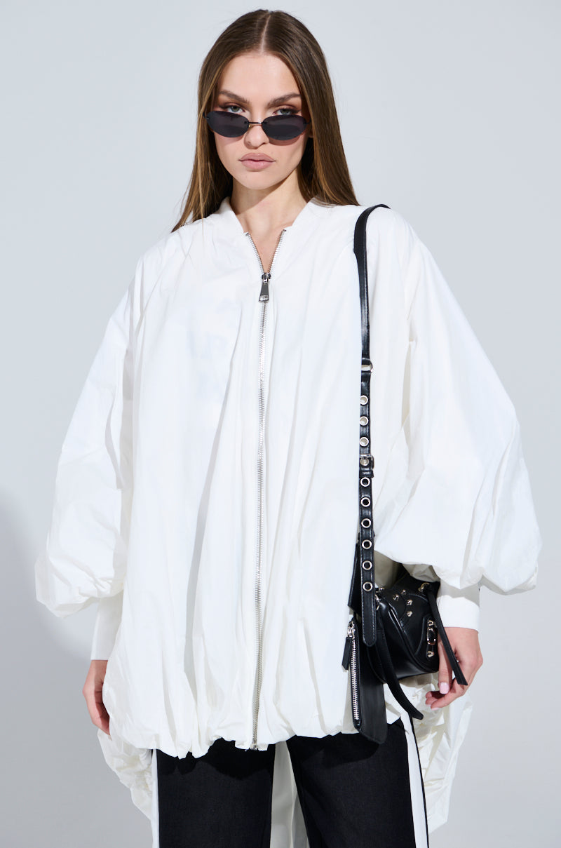 A person in a white FLOATING NYLON BUBBLE BOMBER with a ribbed collar, black sunglasses, and black pants stands against a gray background. They carry a black bag with metal details on their shoulder, and their long, straight hair perfectly complements the oversized silhouette.
