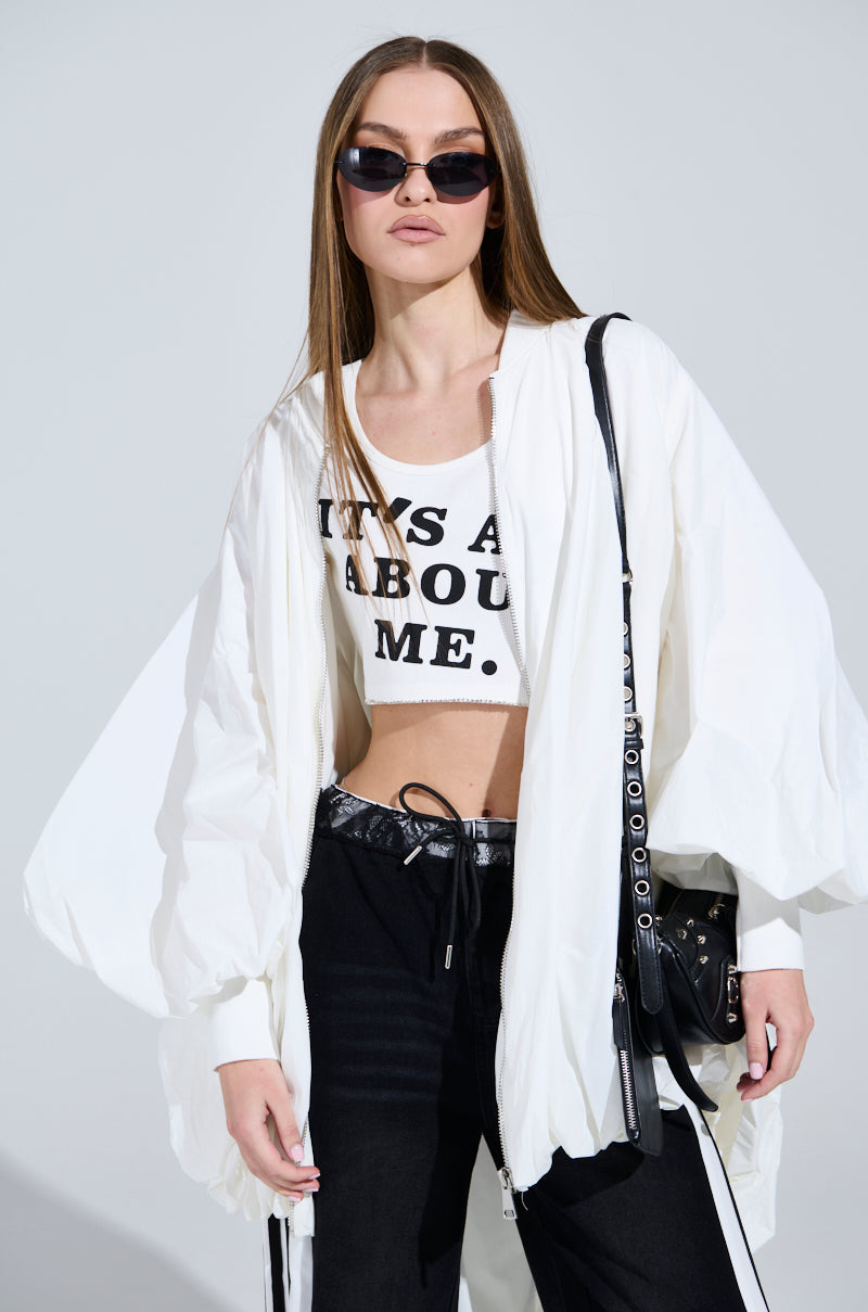 A person with long hair wears sunglasses and an oversized FLOATING NYLON BUBBLE BOMBER with a ribbed collar. Their cropped top has partially visible text, paired with black pants featuring a white stripe. They carry a black bag, standing confidently against a plain background.