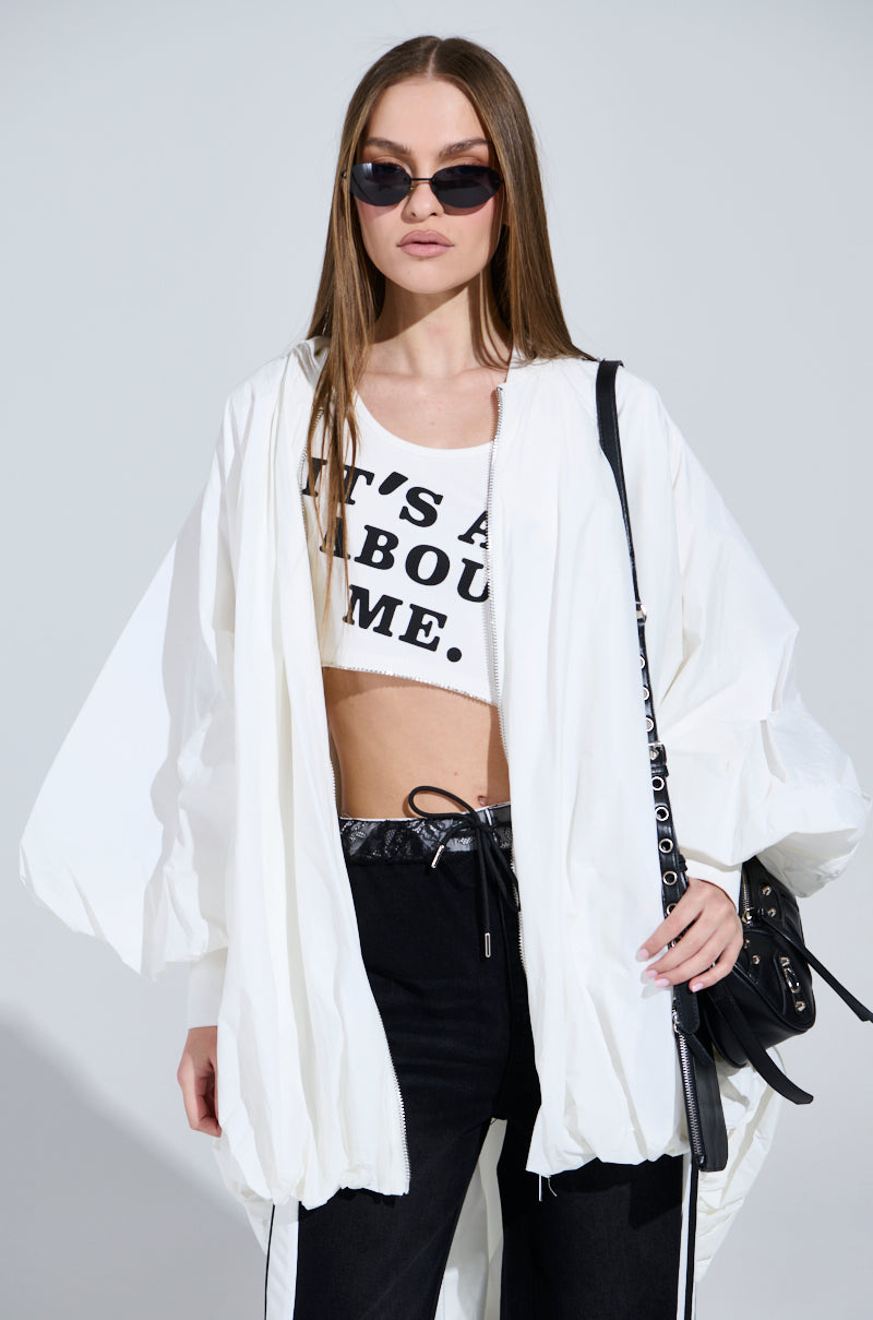 Sporting an oversized silhouette in a white FLOATING NYLON BUBBLE BOMBER with a ribbed collar, a person with long hair and black sunglasses pairs it with a bold crop top and black pants, holding a black bag against a plain background.