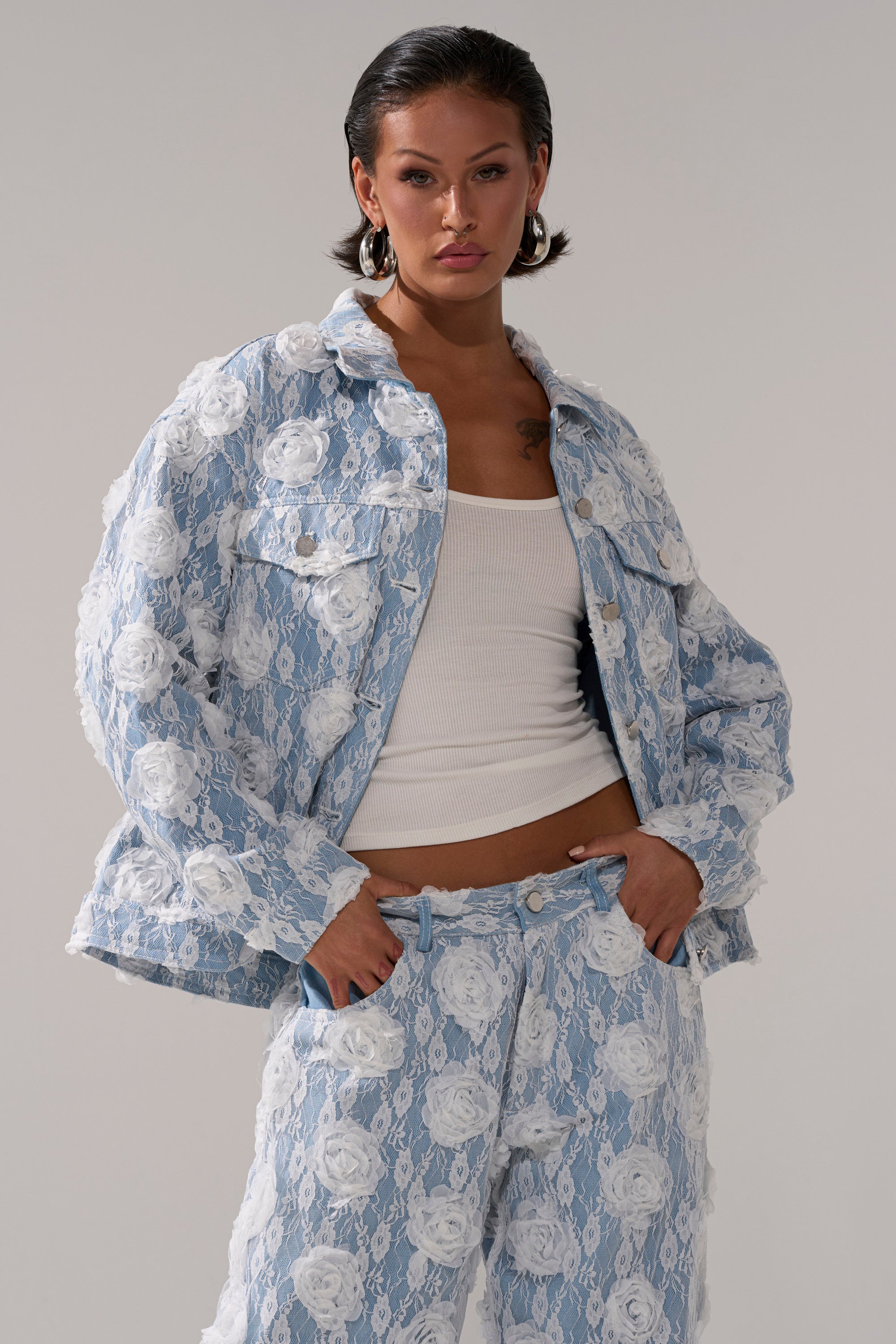 A woman with short dark hair and hoop earrings wears the FOLLOW MY LEAD APPLIQUE DENIM JACKET and matching pants featuring light blue denim with white floral designs, styled over a white ribbed tank top. She poses with her hands in her pockets.