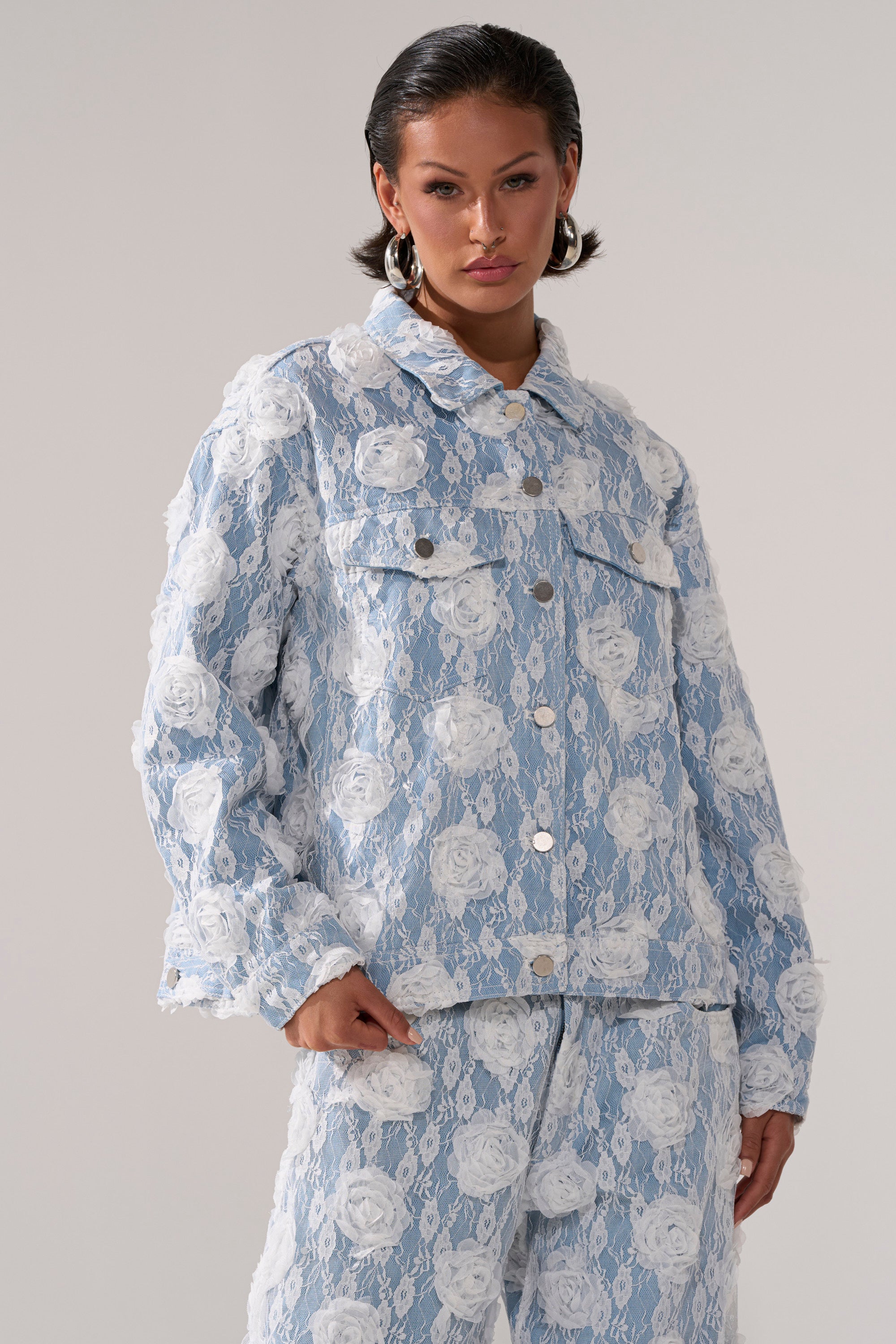A woman wears the FOLLOW MY LEAD APPLIQUE DENIM JACKET and matching pants with white floral lace, standing against a plain background, her hair slicked back and accessorized with large hoop earrings.