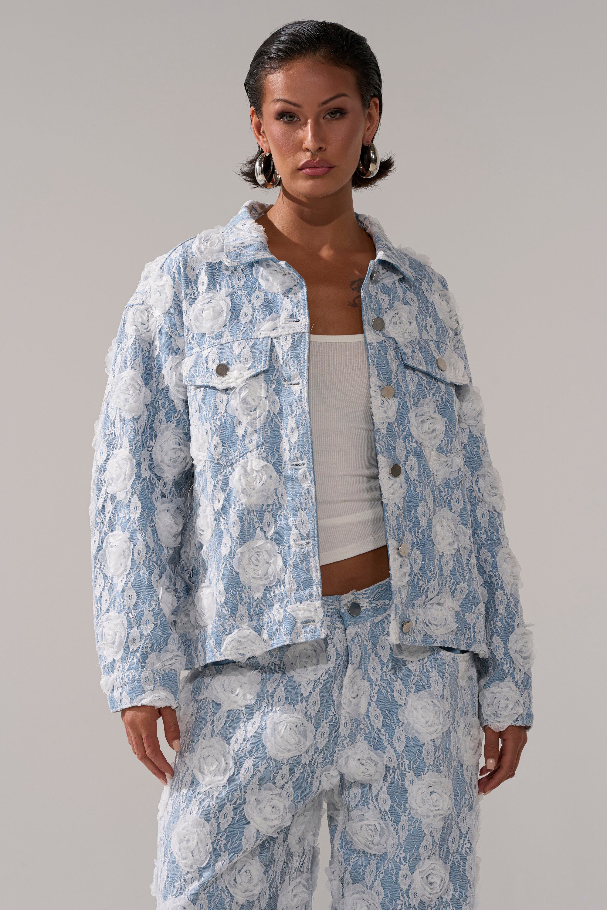 Wearing the FOLLOW MY LEAD APPLIQUE DENIM JACKET with matching pants and a white floral lace overlay, paired with a white ribbed tank, a person poses against a plain background.