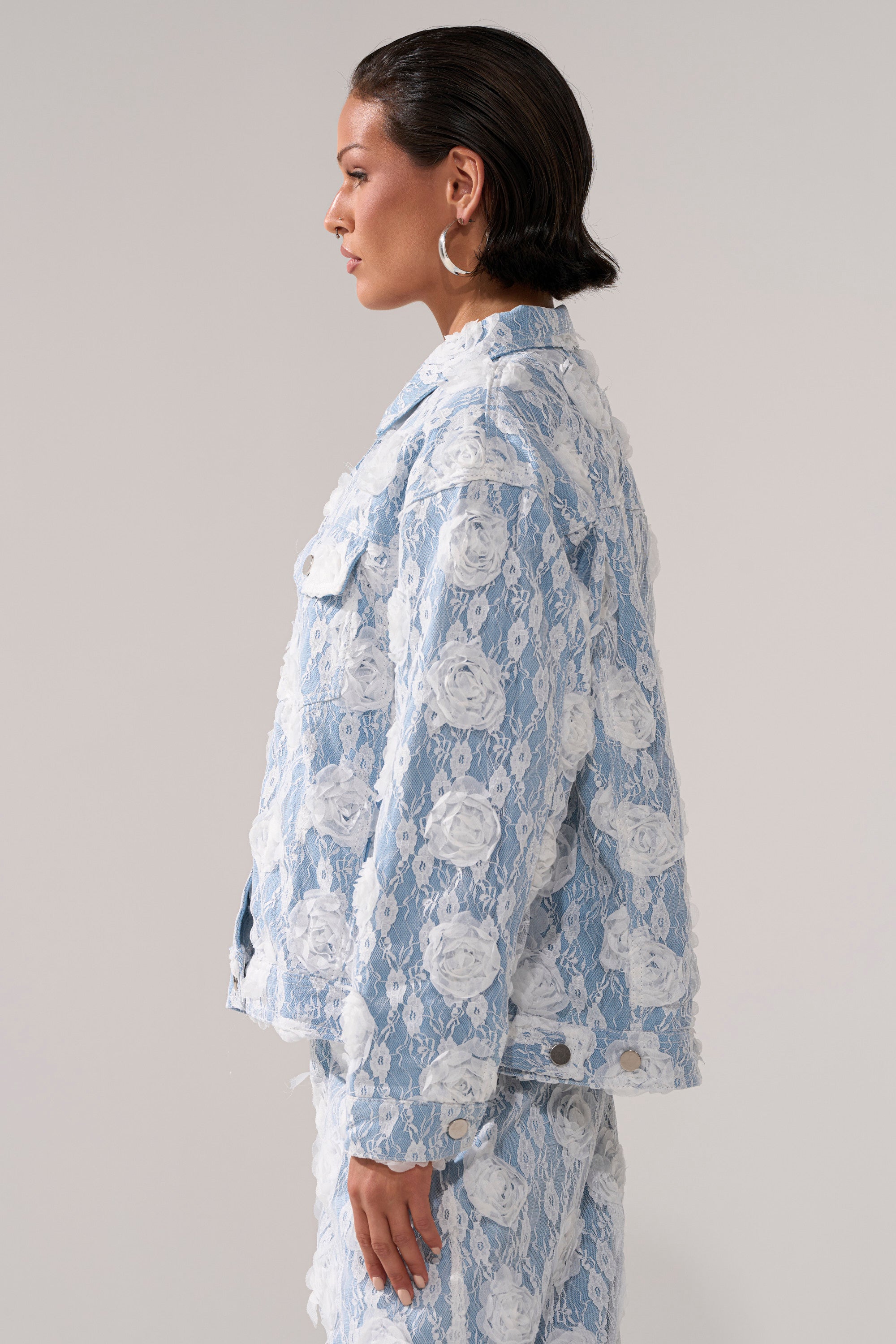 A woman with short dark hair and large hoop earrings stands in profile, wearing the FOLLOW MY LEAD APPLIQUE DENIM JACKET—blue and white denim with floral lace overlay—paired with matching pants against a plain light background.