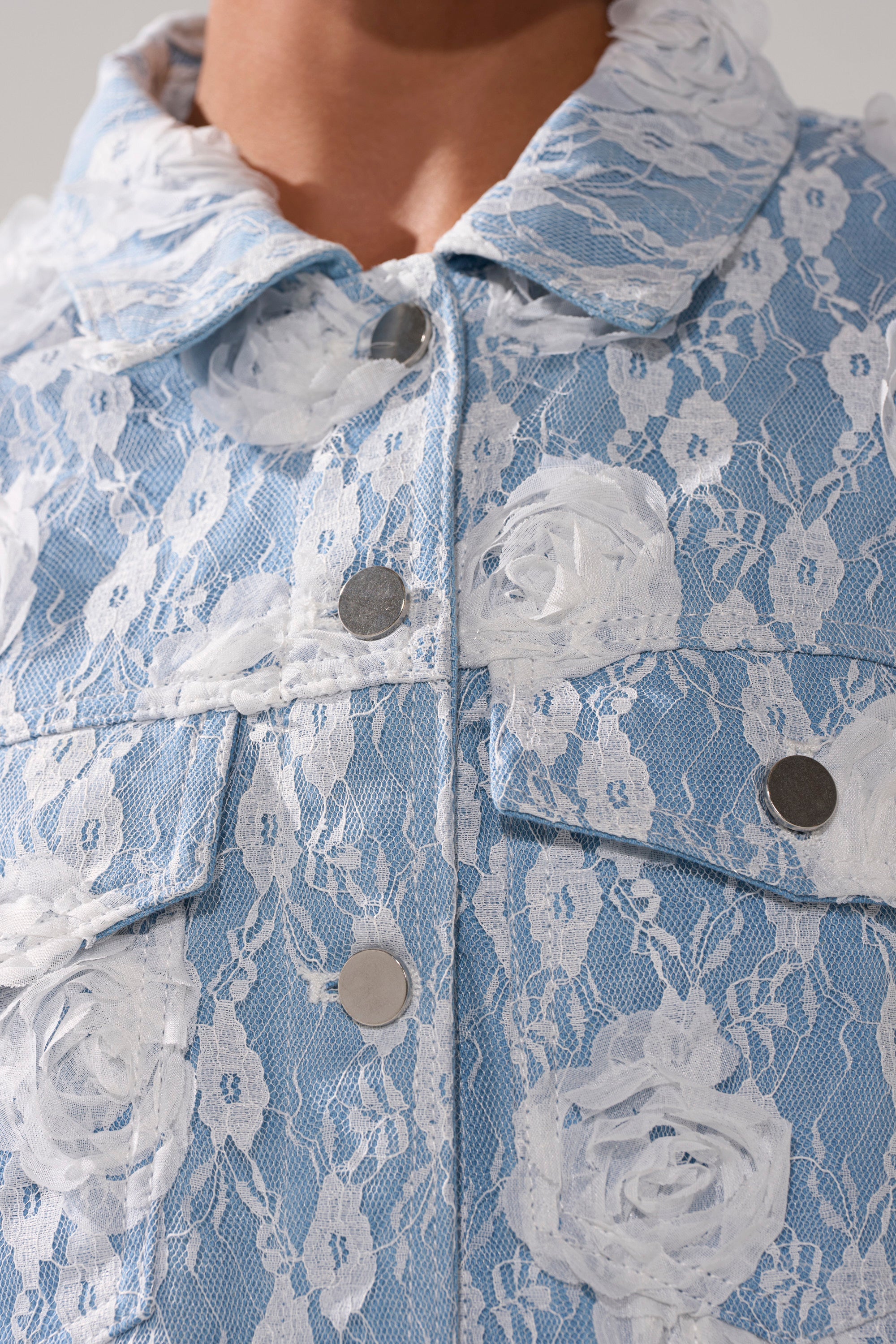 Close-up of the FOLLOW MY LEAD APPLIQUE DENIM JACKET, an oversized denim jacket with white rose-patterned lace applique, metallic snap buttons, and a collar for a unique textured look.