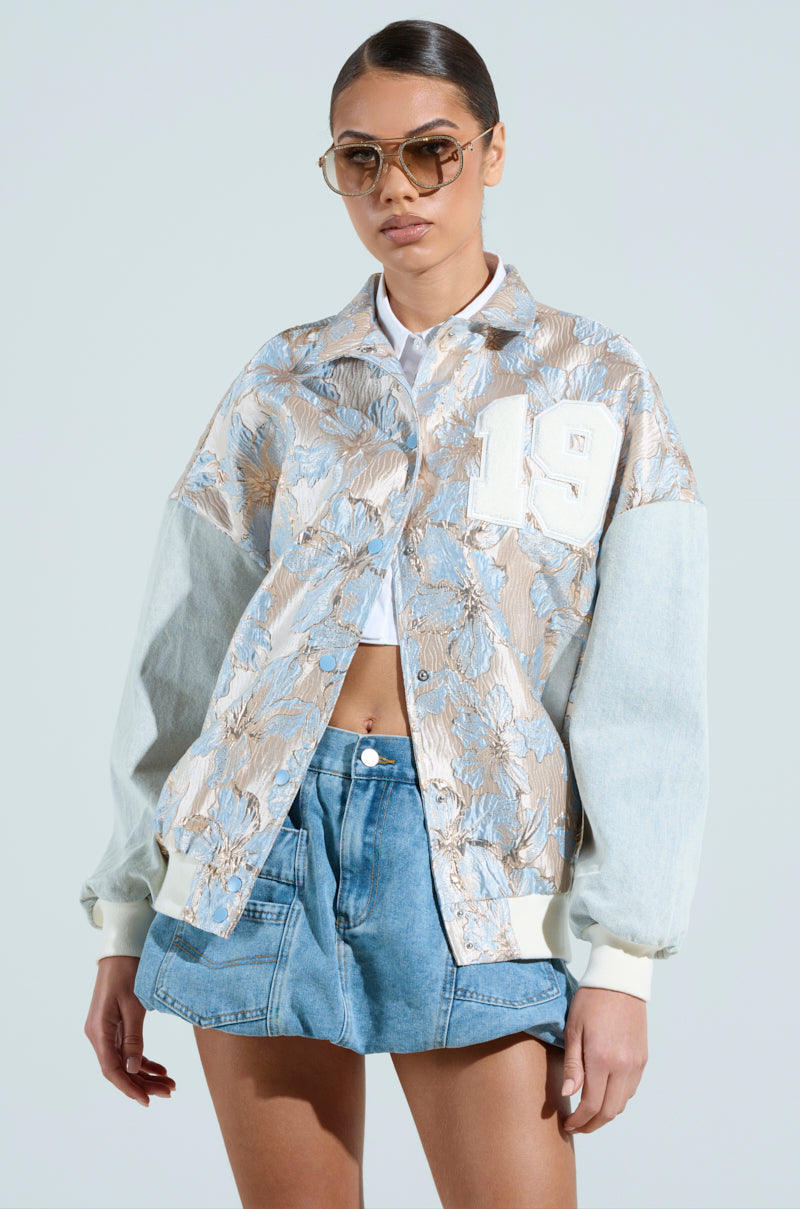 Wearing the ALIYA BROCADE BOMBER with denim sleeves and a metallic floral pattern featuring the number 19, a person pairs it with sunglasses, a white crop top, and high-waisted denim shorts against a plain background, exuding confidence and style.