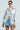 Wearing the ALIYA BROCADE BOMBER with denim sleeves and a metallic floral pattern featuring the number 19, a person pairs it with sunglasses, a white crop top, and high-waisted denim shorts against a plain background, exuding confidence and style.