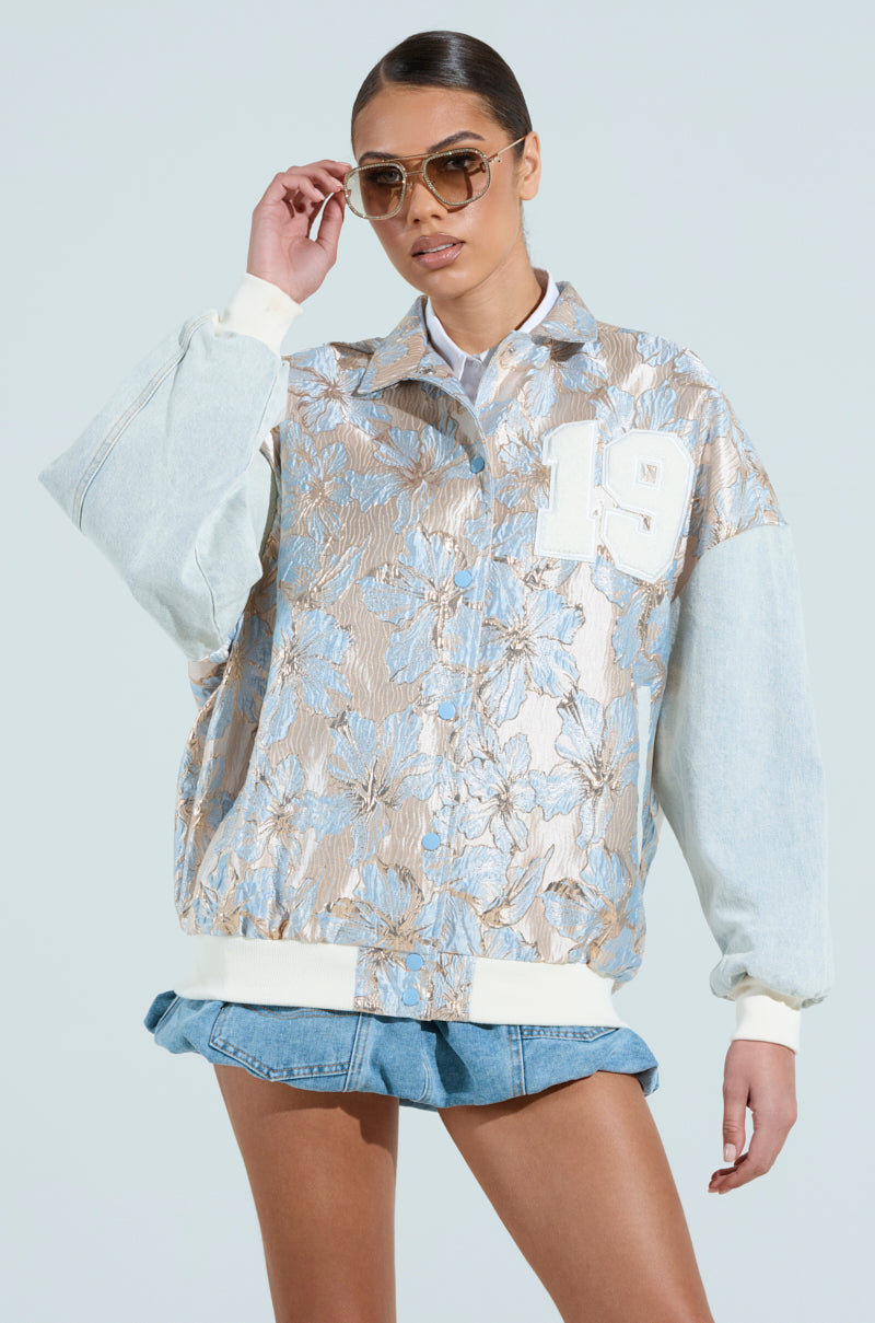 A person wearing sunglasses poses in the ALIYA BROCADE BOMBER, featuring a metallic floral design and a white "19" patch. They also wear a light blue denim mini skirt against a solid light blue background.