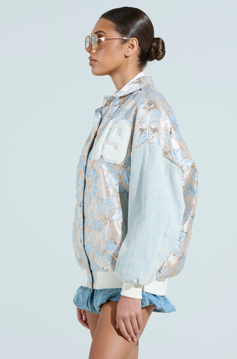 Wearing the ALIYA BROCADE BOMBER with its chic metallic floral pattern, a person in denim shorts stands in profile against a light grey background. Their look is completed with oversized sunglasses and neatly pulled-back hair.