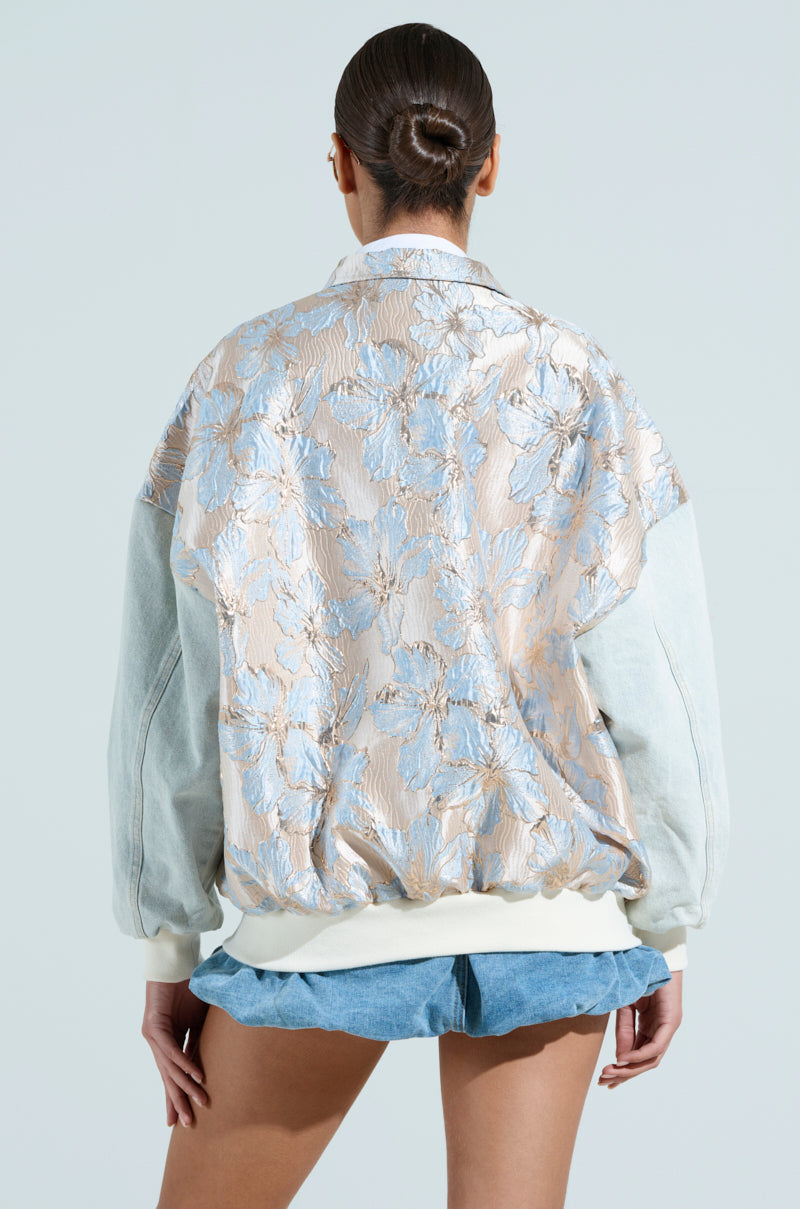 A person is seen from behind wearing an ALIYA BROCADE BOMBER with a floral pattern in light blue and beige tones, featuring wide sleeves and a loose fit. They pair it with a light blue skirt, and their hair is styled in a bun.