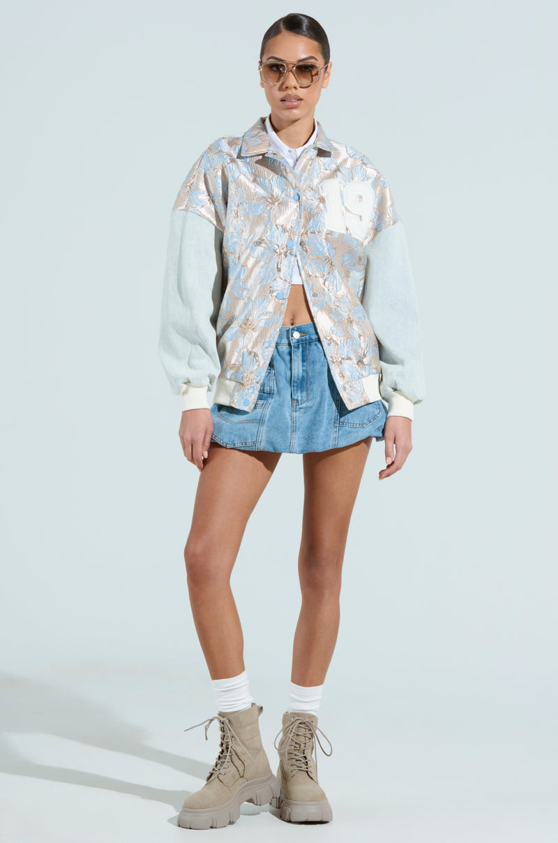 A person stands confidently in an ALIYA BROCADE BOMBER with metallic floral patterns, a denim mini skirt, boots, thick socks, and sunglasses against a light-colored background.